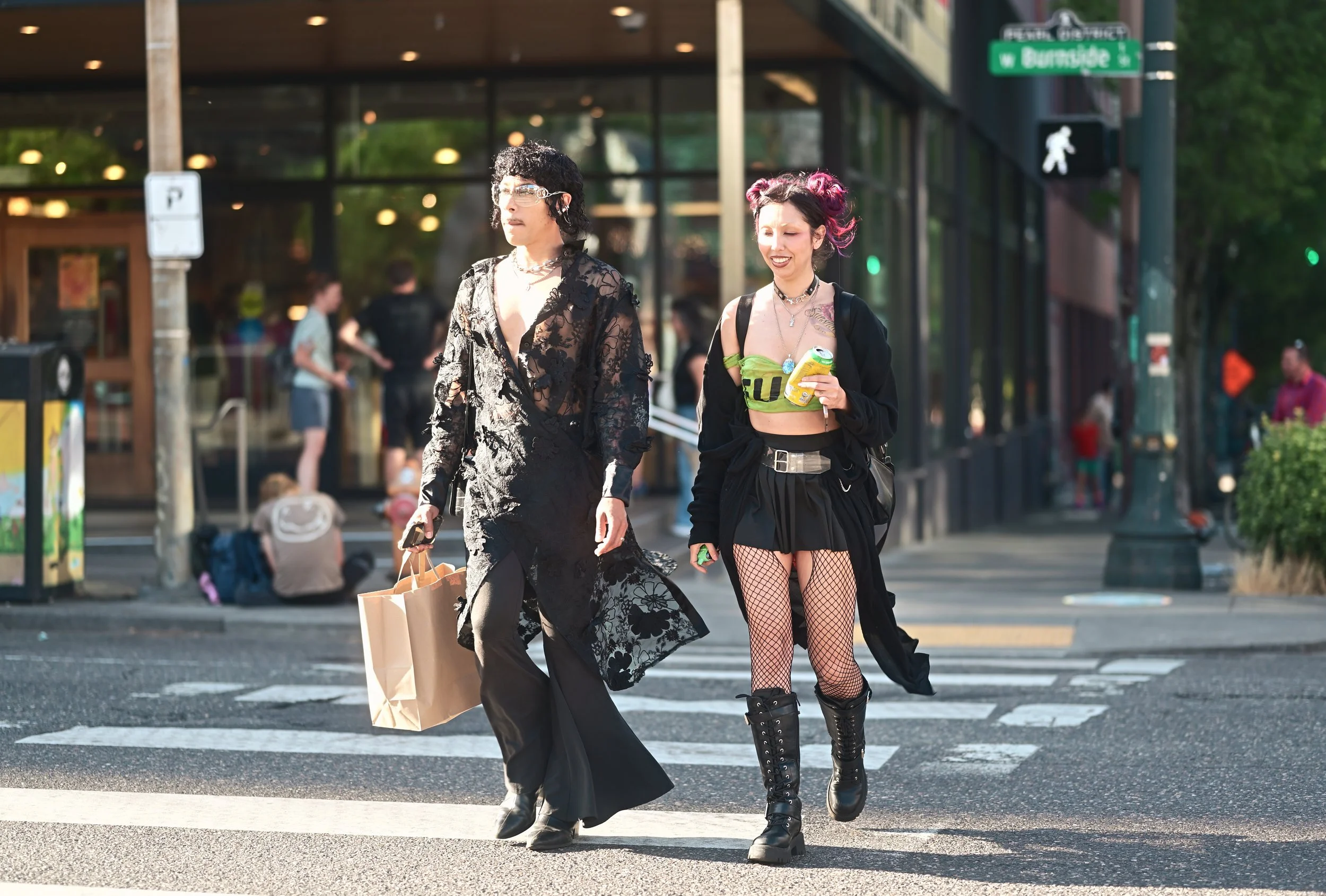 Two women walking across a crosswalk in an urban area, dressed in alternative fashion with one wearing a black lace dress and the other in a black outfit with fishnet stockings and combat boots, carrying shopping bags and holding drinks.