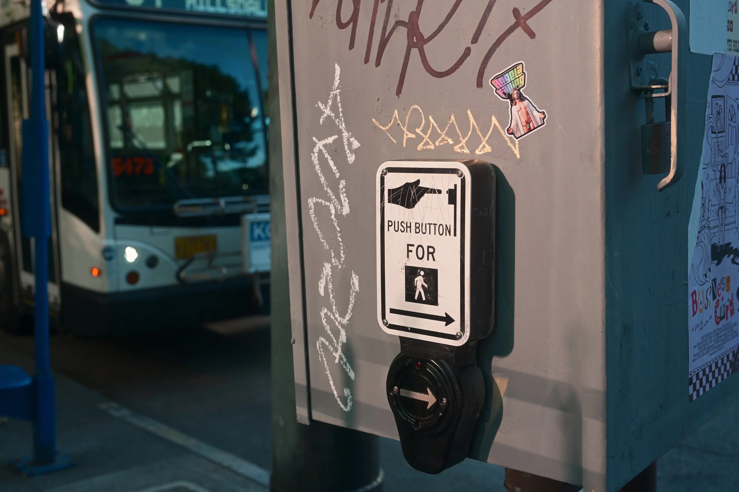 A walk button station on a city street, with a sticker of a person and colorful text, and a bus in the background.