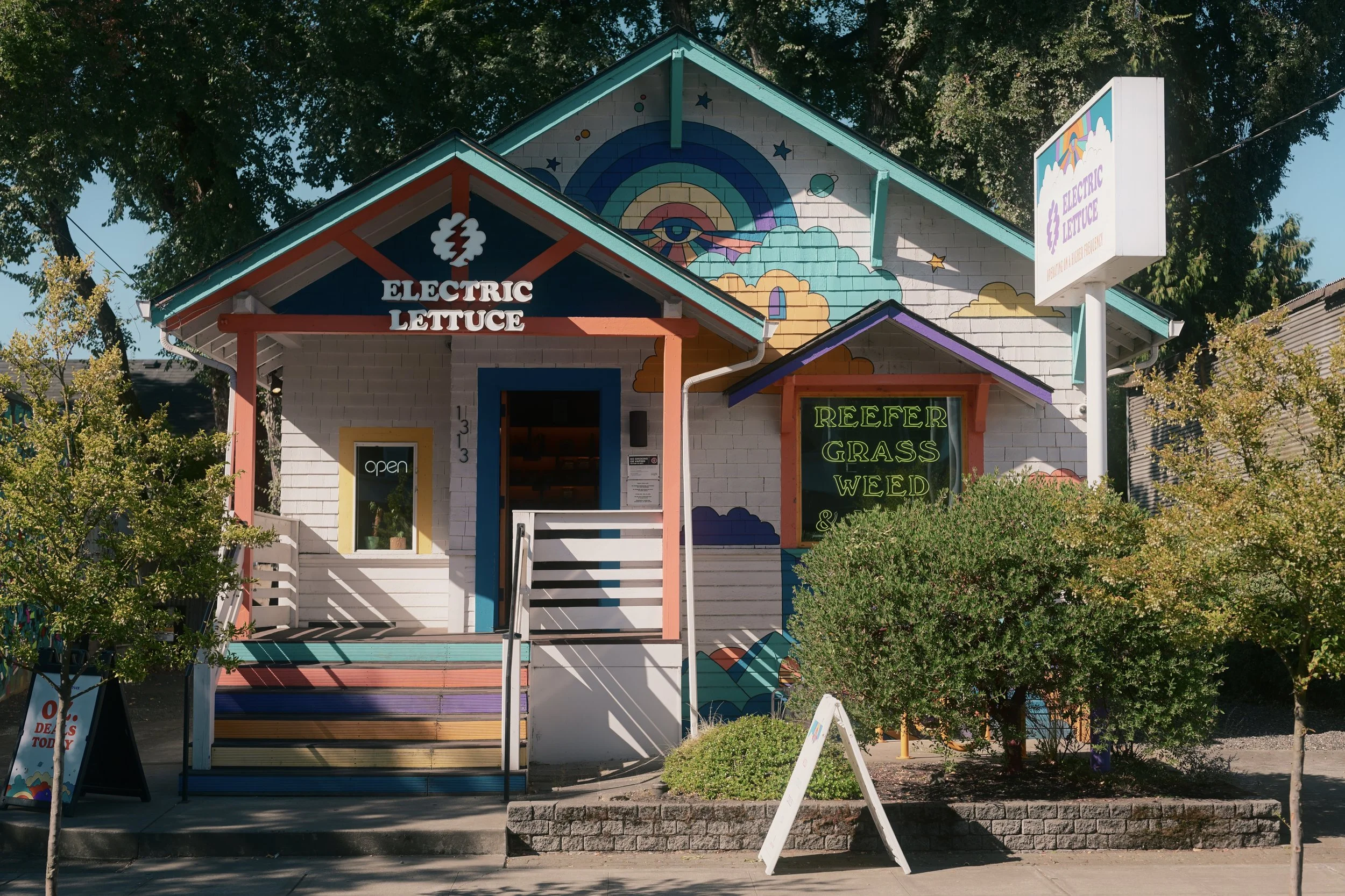 Colorful building with a sign reading 'Electric Lettuce' and a neon sign advertising 'Reefer Grass Weed'. Bright mural of an eye, rainbow, birds, and clouds painted on the upper part of the building. Surrounded by trees and other greenery.