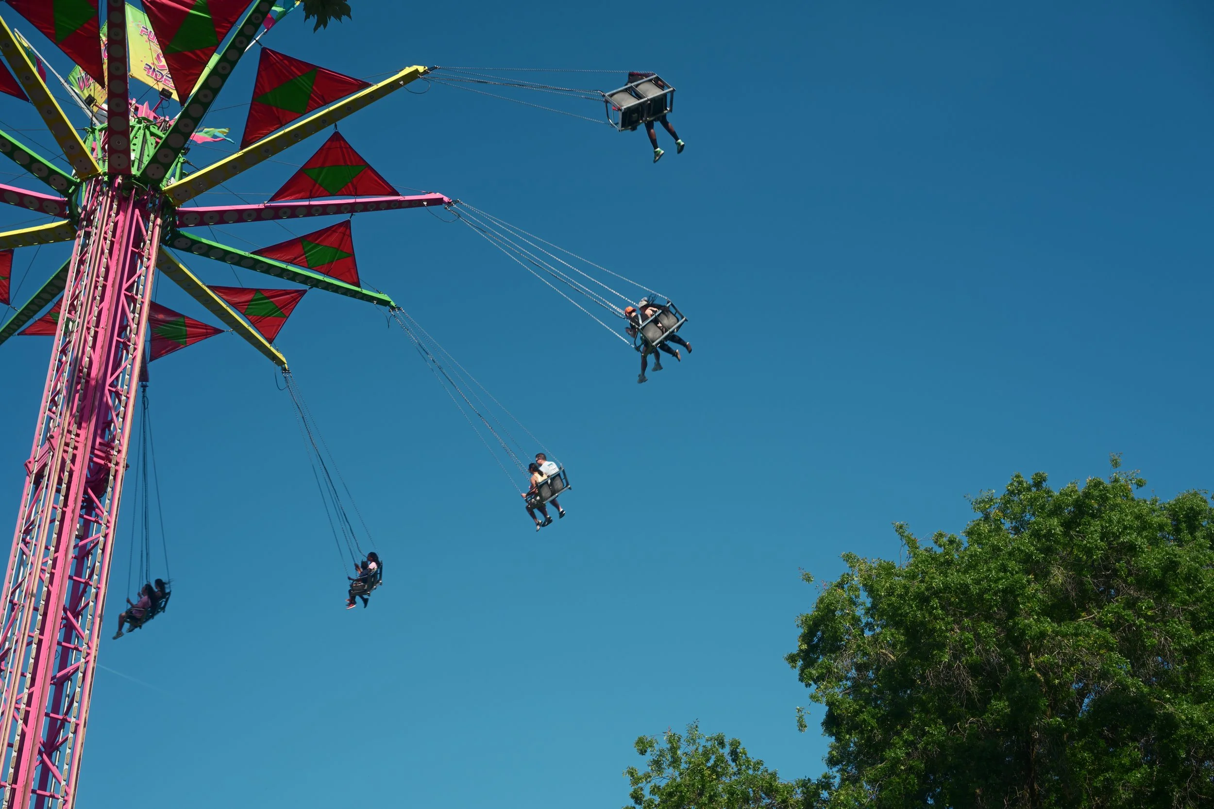 People riding on swings of a large colorful amusement park ride against a clear blue sky with some trees at the bottom right corner.