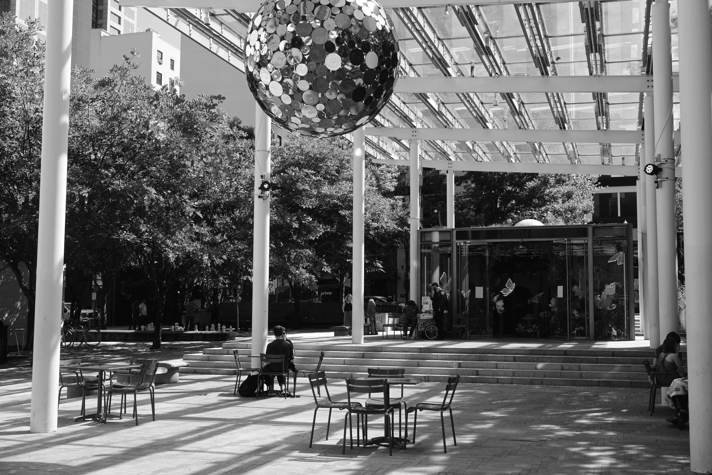 Outdoor public space with tables and chairs, people sitting and walking, trees, and a modern glass and metal structure suspended above with a large spherical sculpture made of reflective circular discs