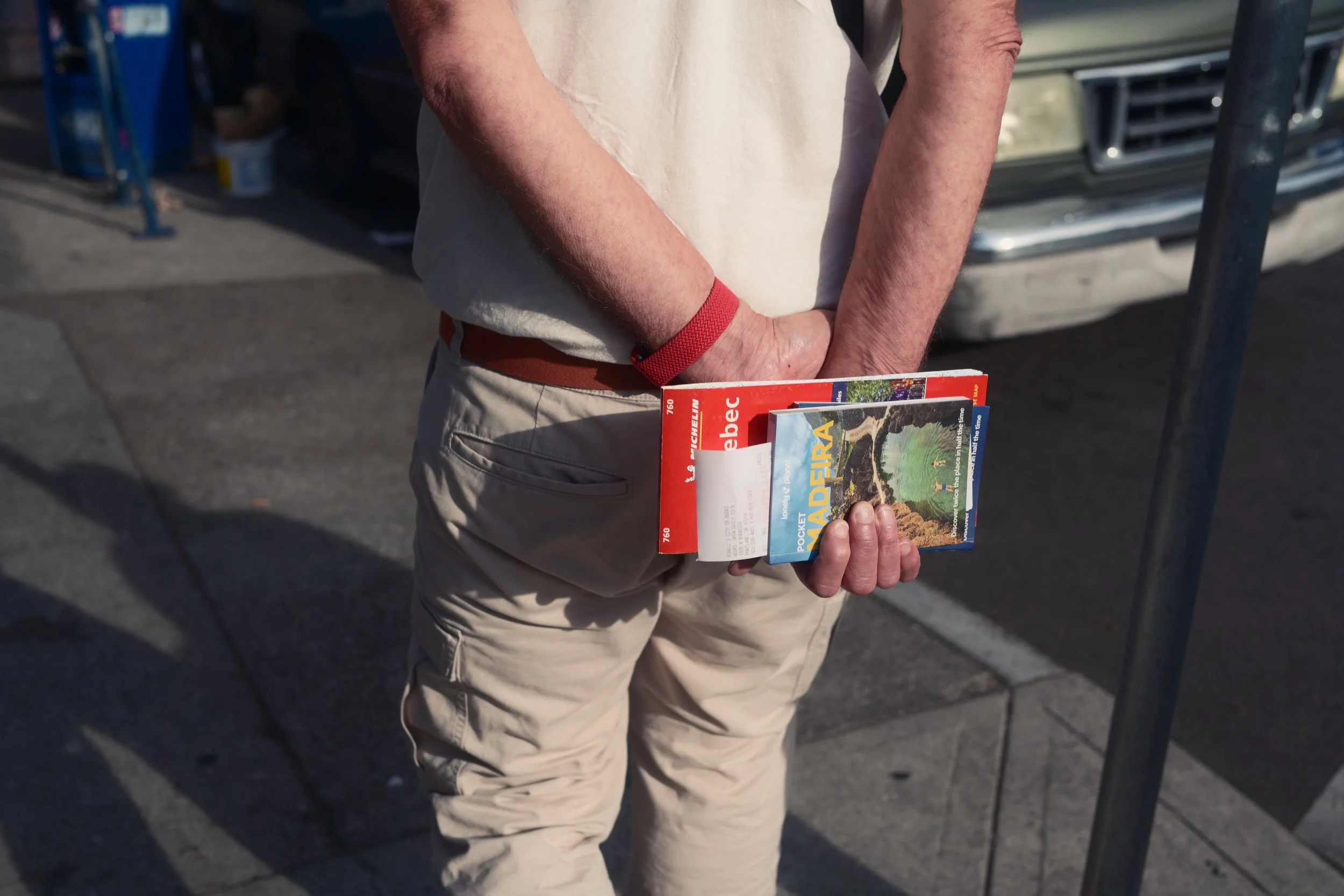 A person holding brochures or maps, standing outdoors on a sidewalk, with a car and some equipment visible in the background.