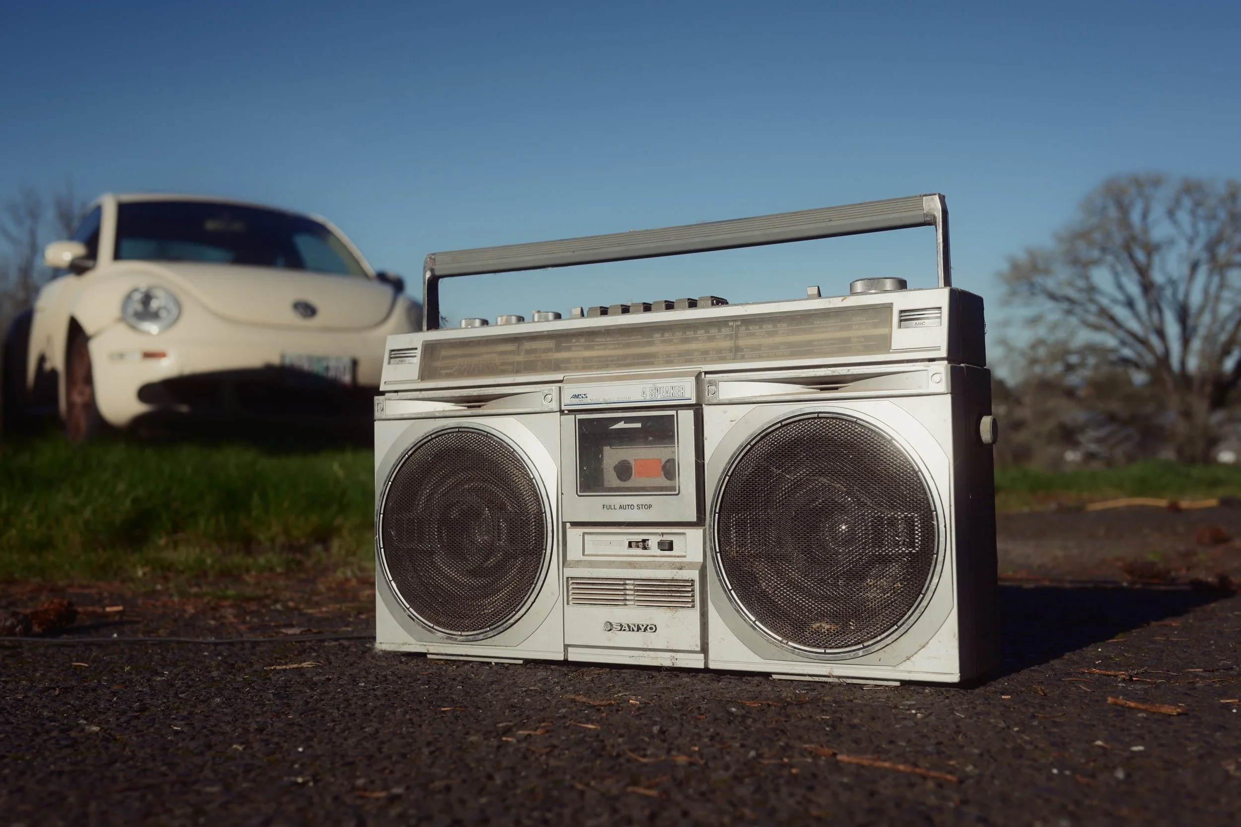 A vintage Sanyo stereo boombox with two speakers and a radio tuner, placed on the ground outdoors. In the background, there is a white Volkswagen Beetle car parked on grass with a clear blue sky overhead.