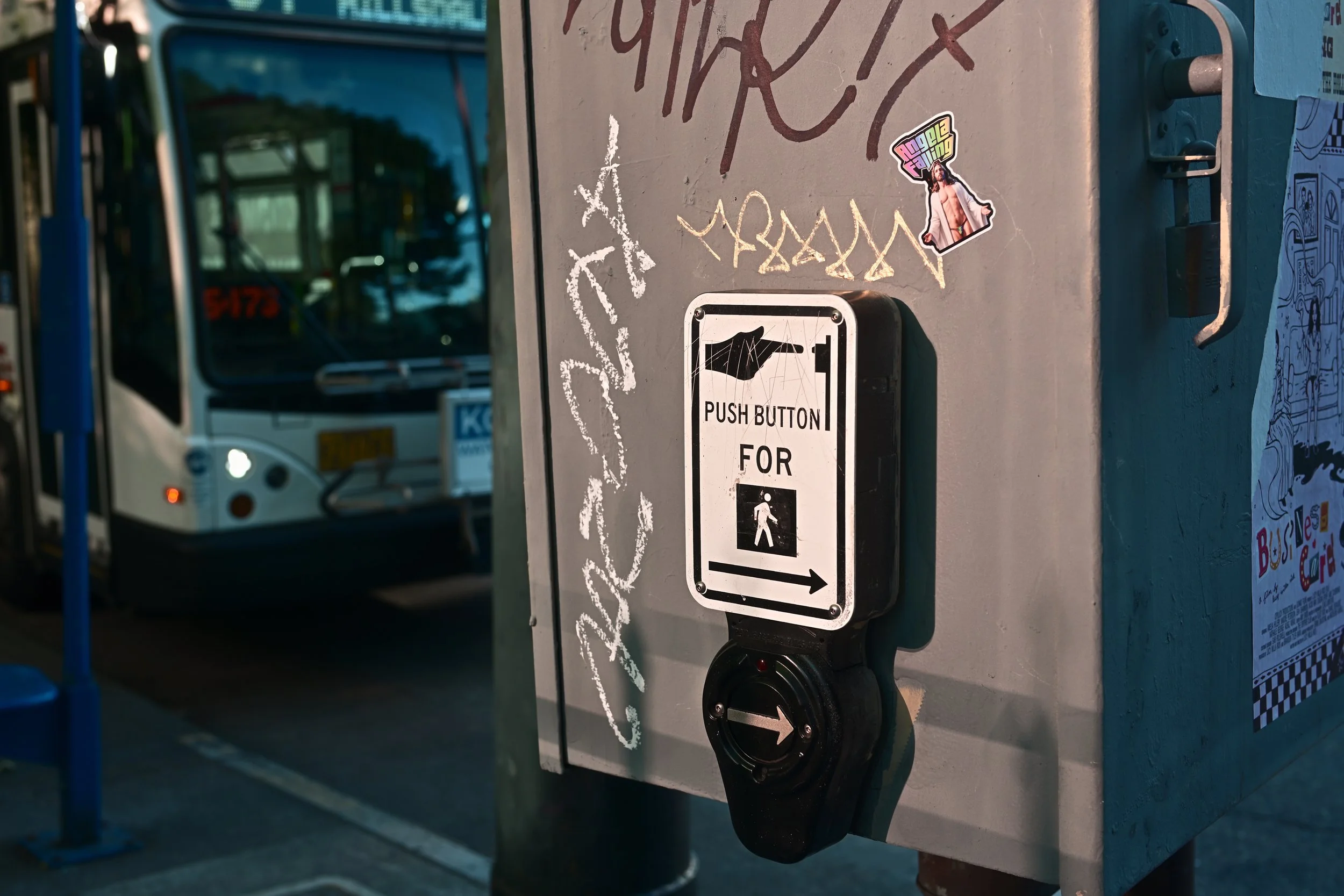 Close-up of a pedestrian push button with a black button and a white sign featuring a black hand icon, indicating to press for crossing, mounted on a gray metal box on a city sidewalk.