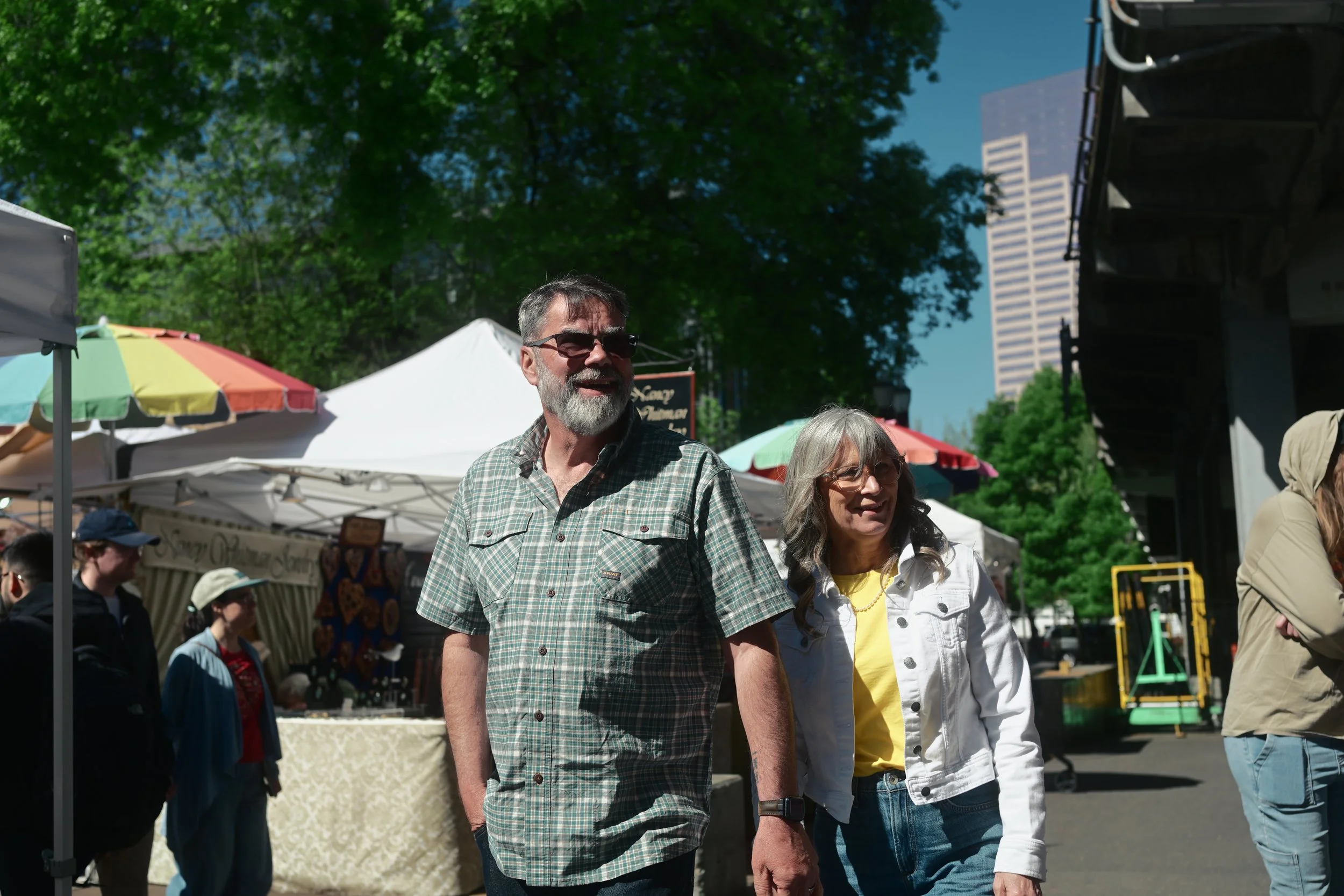 People walking at an outdoor event with tents and trees in the background on a sunny day.