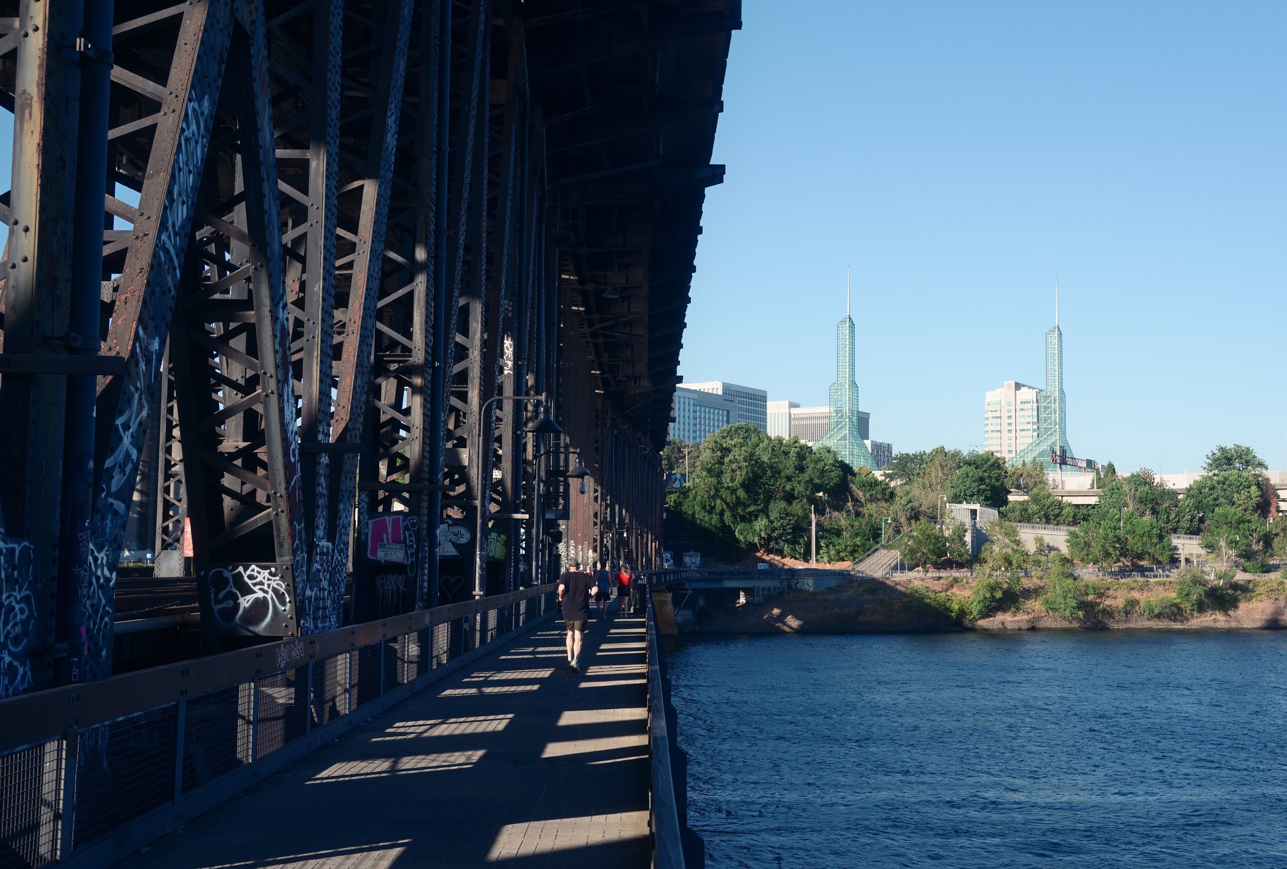 People walking on a bridge over a river in an urban area with tall buildings and green trees in the background, under a clear blue sky.