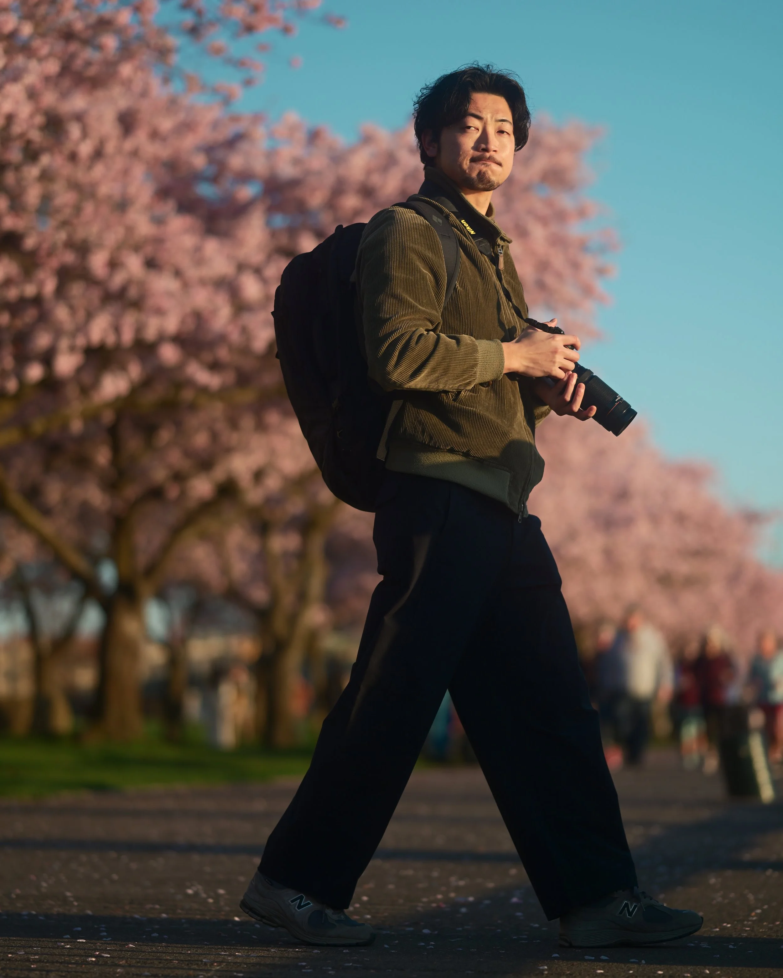 A man wearing dark pants, sneakers, a green jacket, and carrying a camera and backpack walks outdoors past cherry blossom trees in full bloom during late afternoon or early evening.