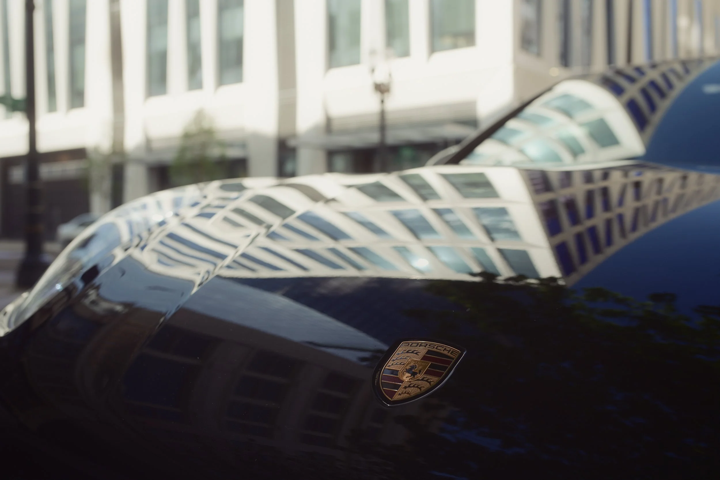 Close-up of a black Porsche vehicle's hood with the Porsche emblem, reflected in nearby glass building windows.