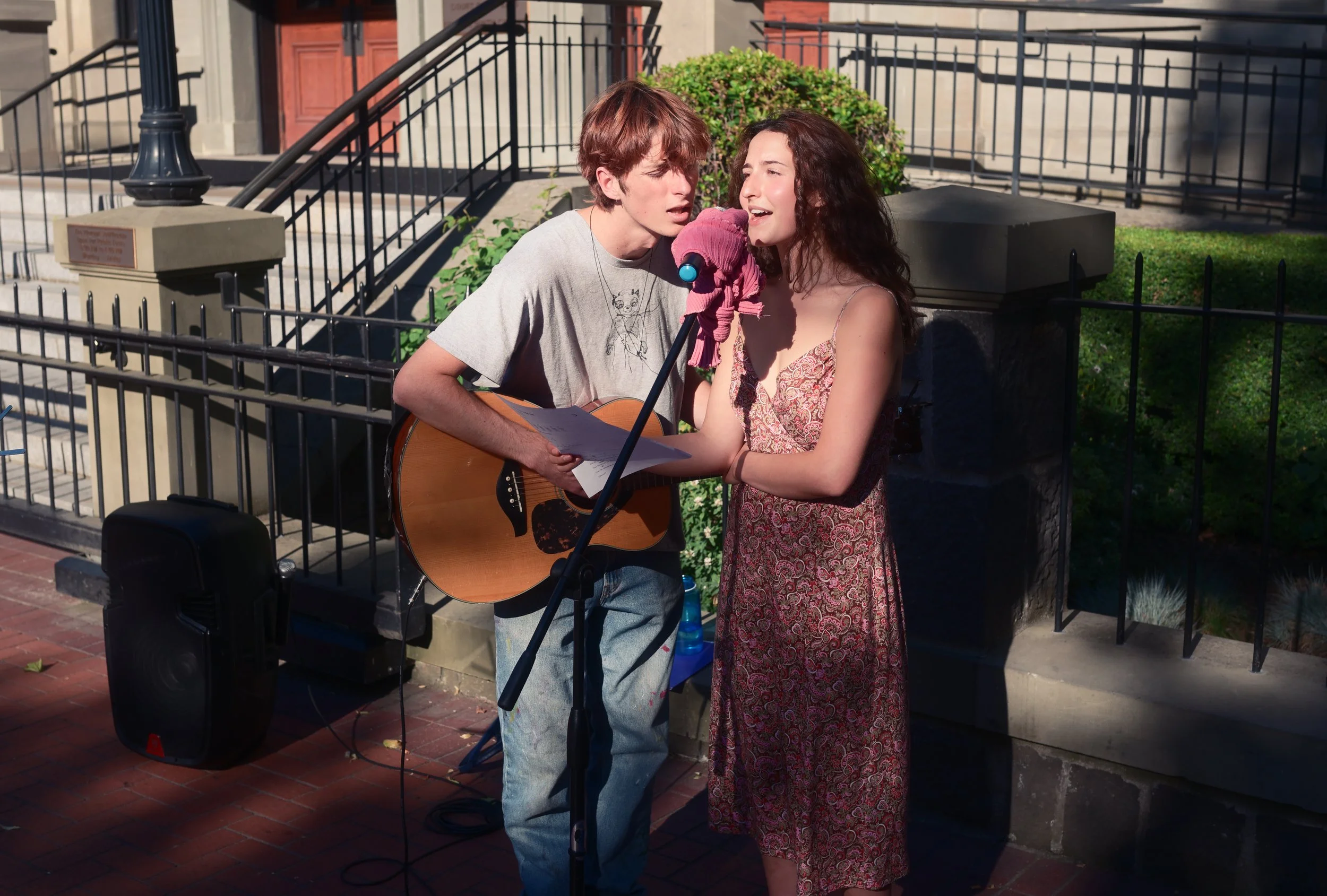 Young man with a guitar and a young woman singing into a pink microphone on a city sidewalk, performing outdoors.