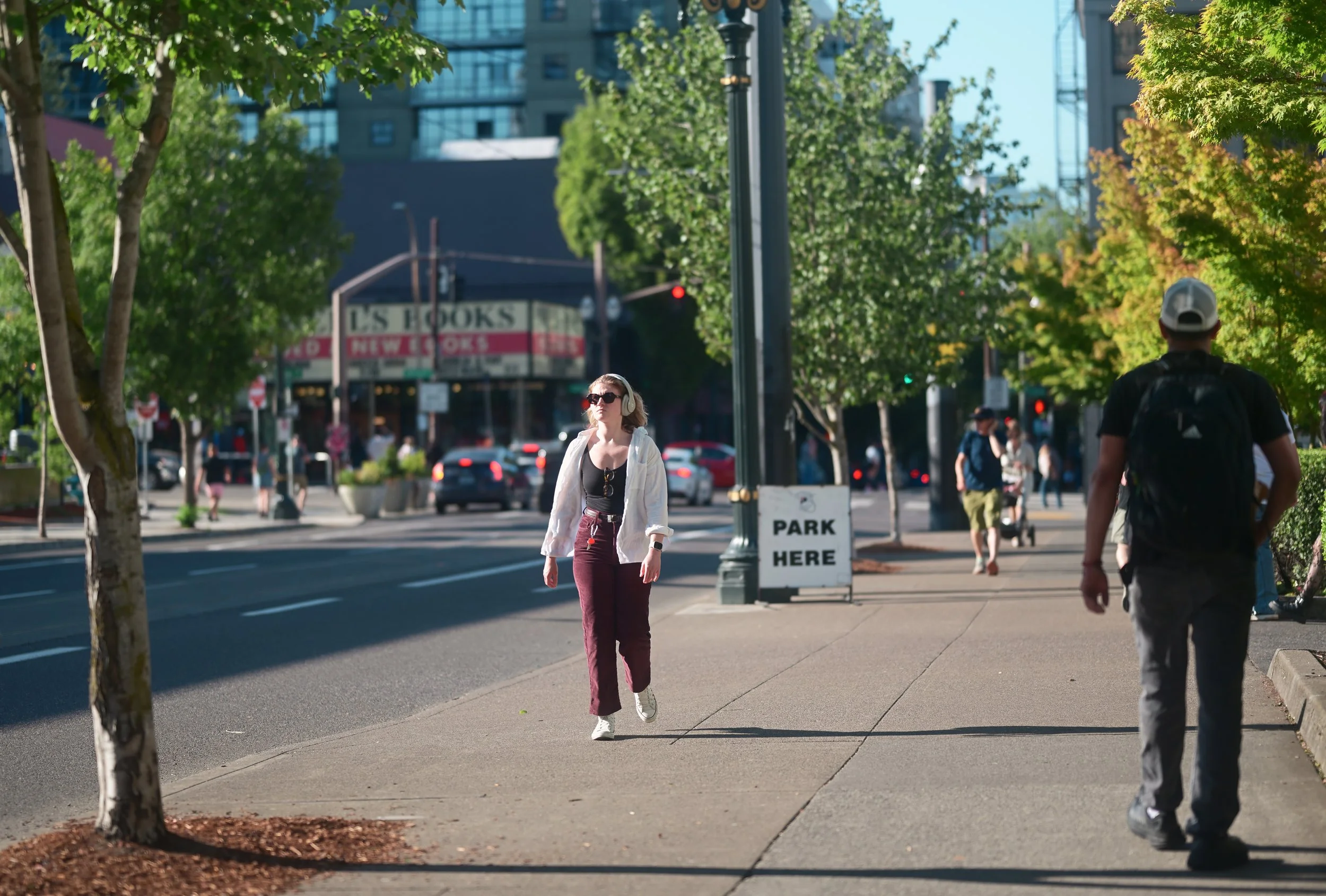 A woman walking on a city sidewalk, wearing sunglasses, headphones, a white jacket, and maroon pants, with trees and buildings in the background.