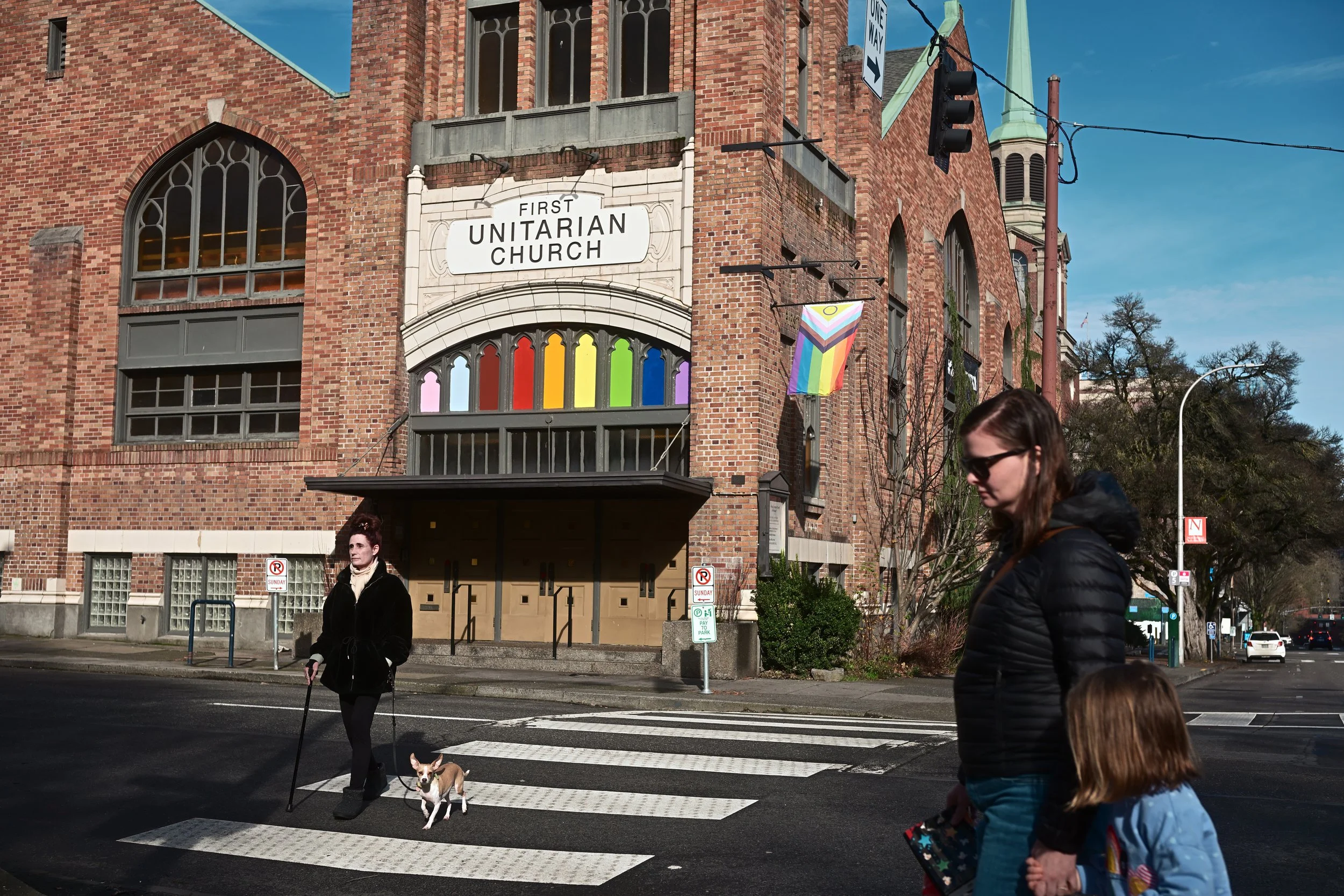 A brick church with stained glass windows and a rainbow pride flag hanging outside. People, including a woman with a dog, are crossing the street at a crosswalk in front of the church on a sunny day.