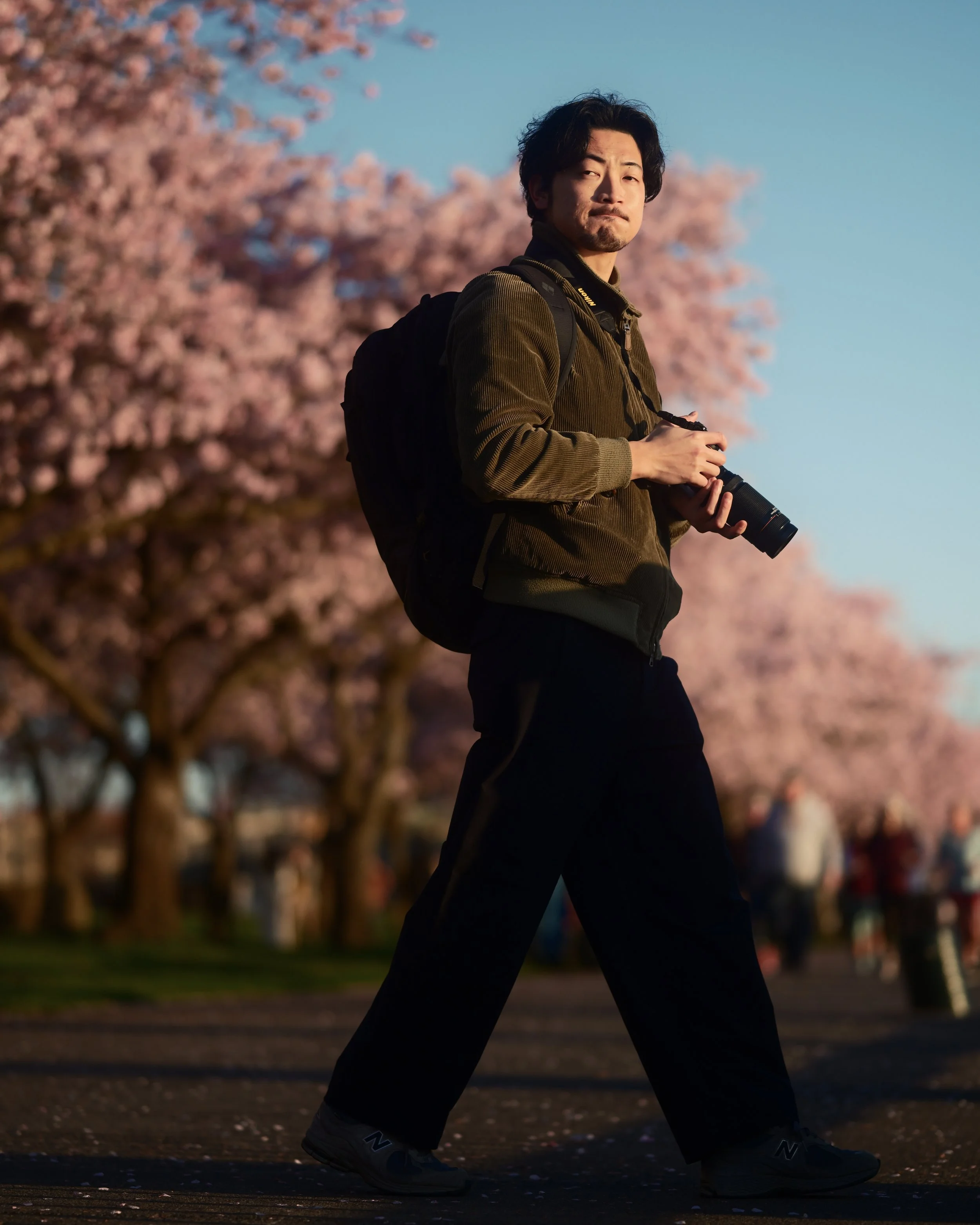 A man walking outdoors with pink flowering trees in the background, holding a camera in his hands and wearing a backpack.