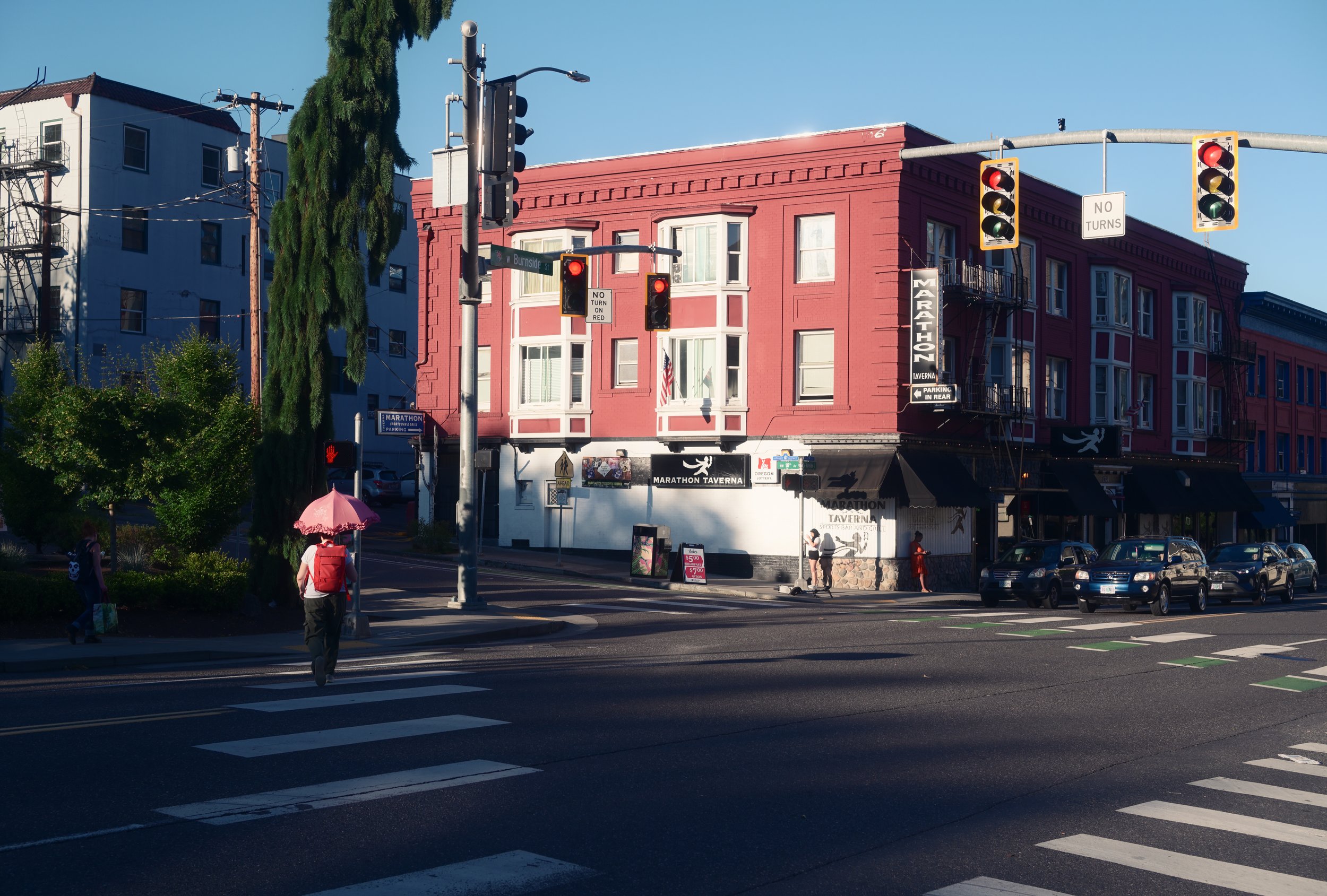 Street corner with traffic lights, pedestrians crossing, and a pink three-story building with signs for Marathon Tavern. Several parked cars and a tree are visible, with clear skies overhead.