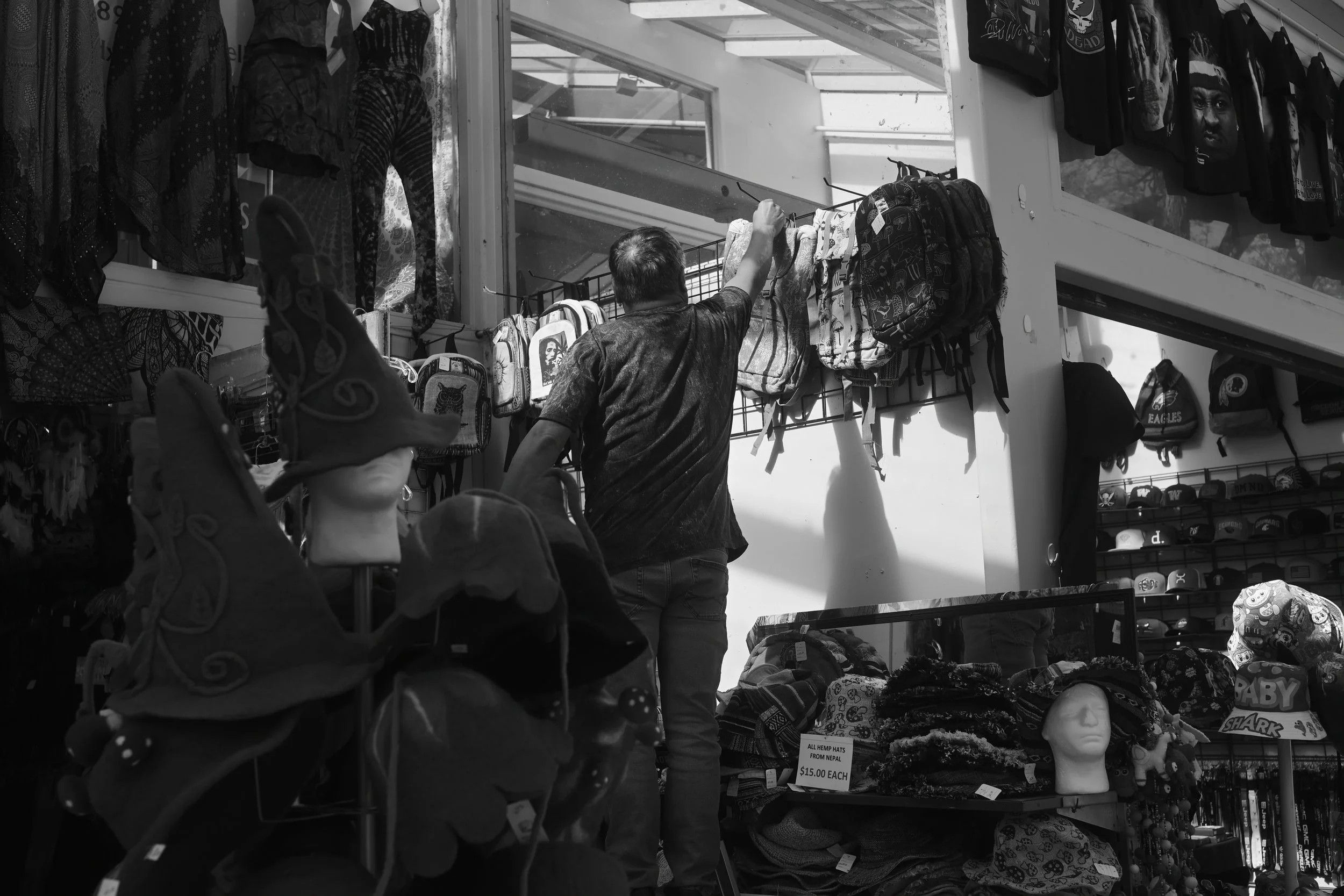 A man shopping at an outdoor market stall with clothing, backpacks, and accessories. The stall's shelves display hats, bags, and shirts, some with graphic designs. The background shows the structure of a building with stairs and windows.