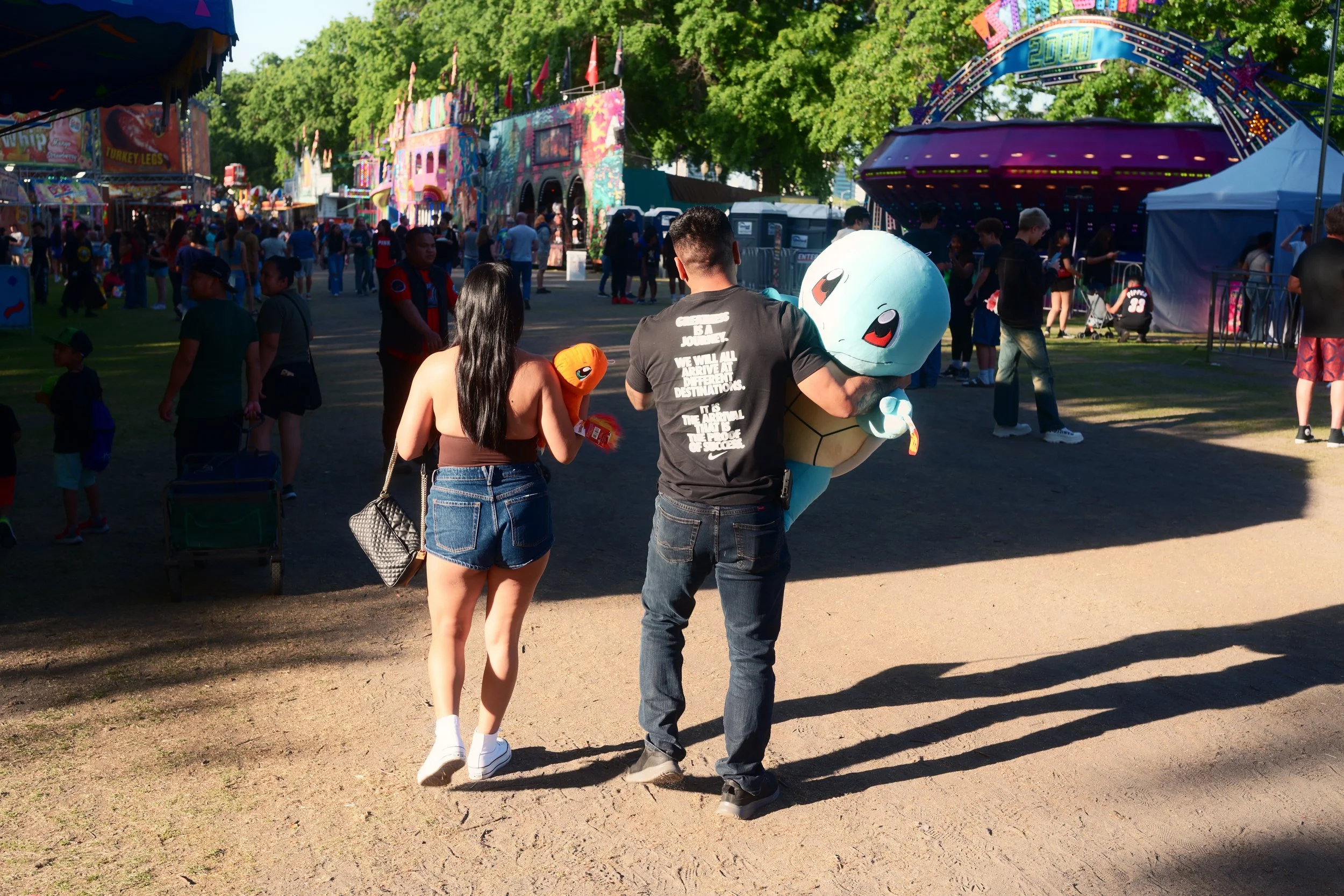 A man and a woman walking through a carnival or fairground, with the man carrying a large Pikachu mascot costume. The background features various amusement rides and booths, with several other people enjoying the event.