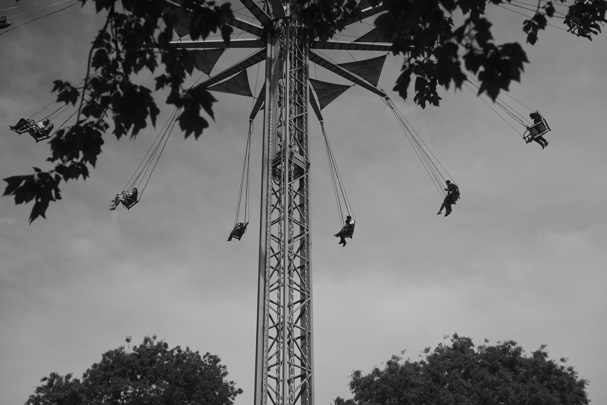 Black and white photo of a tall amusement park ride with people seated on swings hanging from a central tower, with trees at the bottom and a cloudy sky in the background.