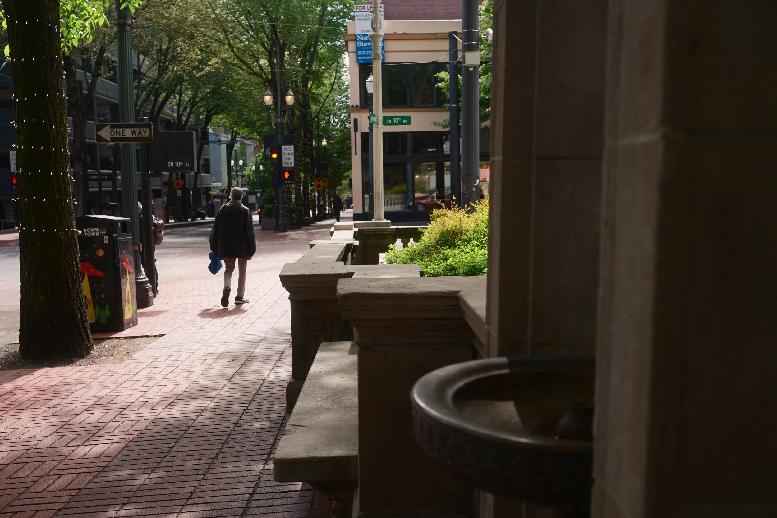 City street scene with trees, pedestrians, and buildings, viewed from under a shelter with a round metal table in the foreground.
