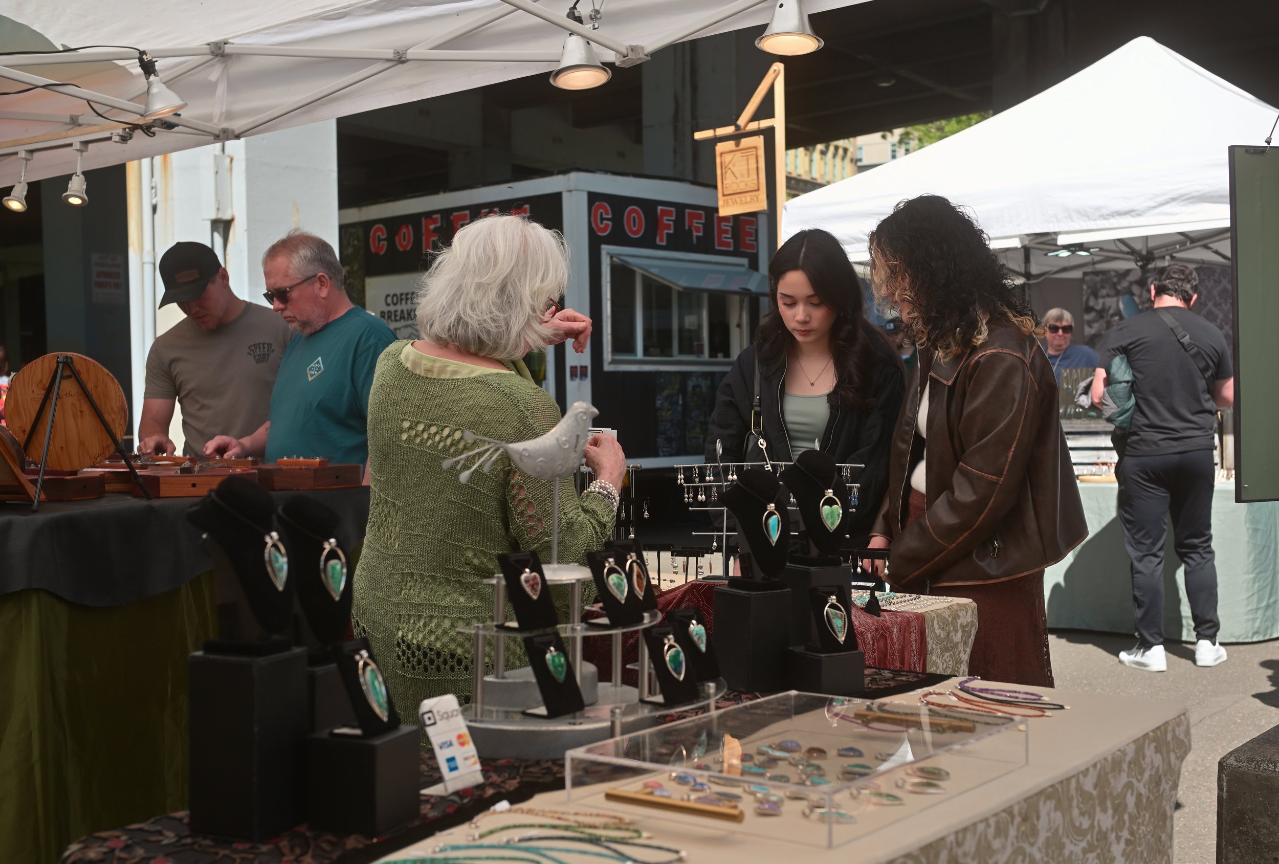 People browsing jewelry and accessories at an outdoor market stall with white tents. The jewelry is displayed on tables and includes necklaces and bracelets with colorful stones and designs.