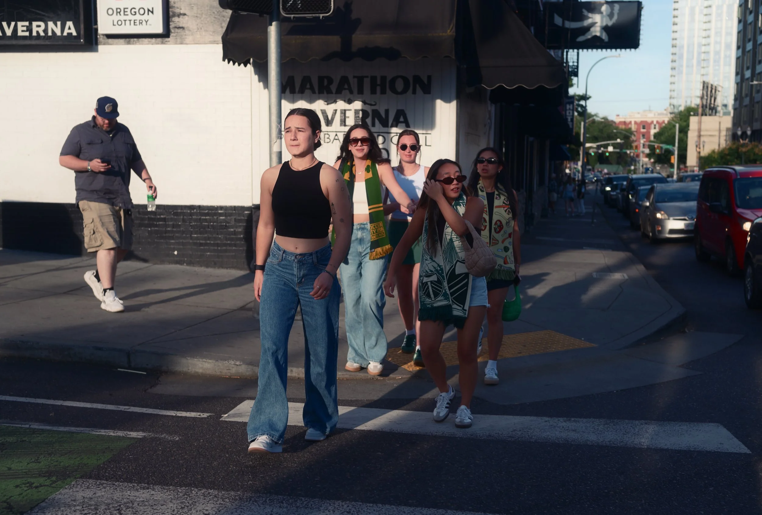 Group of five young women walking on crosswalk, two wearing sunglasses, one in a black crop top with blue jeans, others in casual outfits, city street scene with cars and buildings in the background.