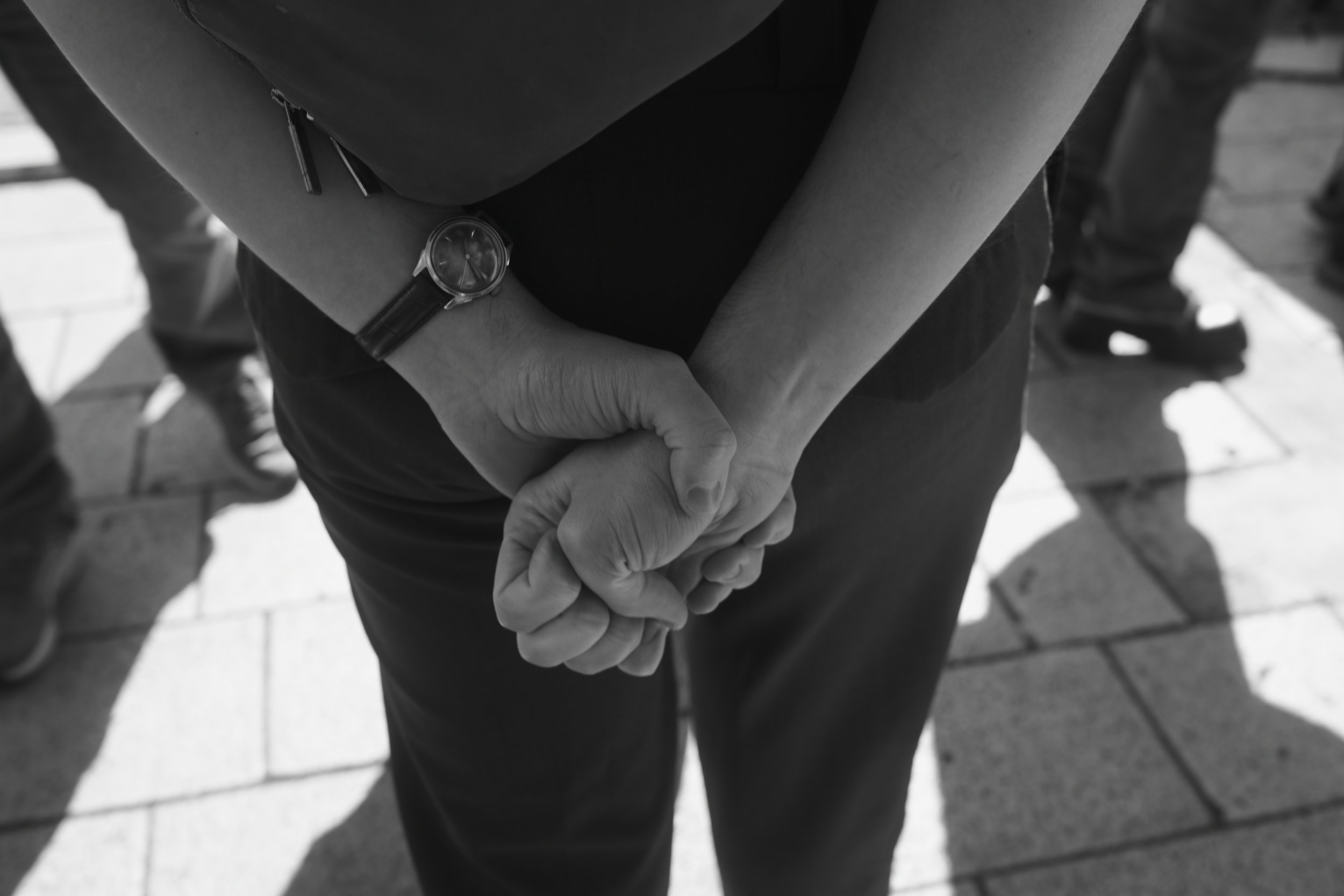 Close-up of two people holding hands in a handshake, with one wearing a wristwatch, on a tiled sidewalk.