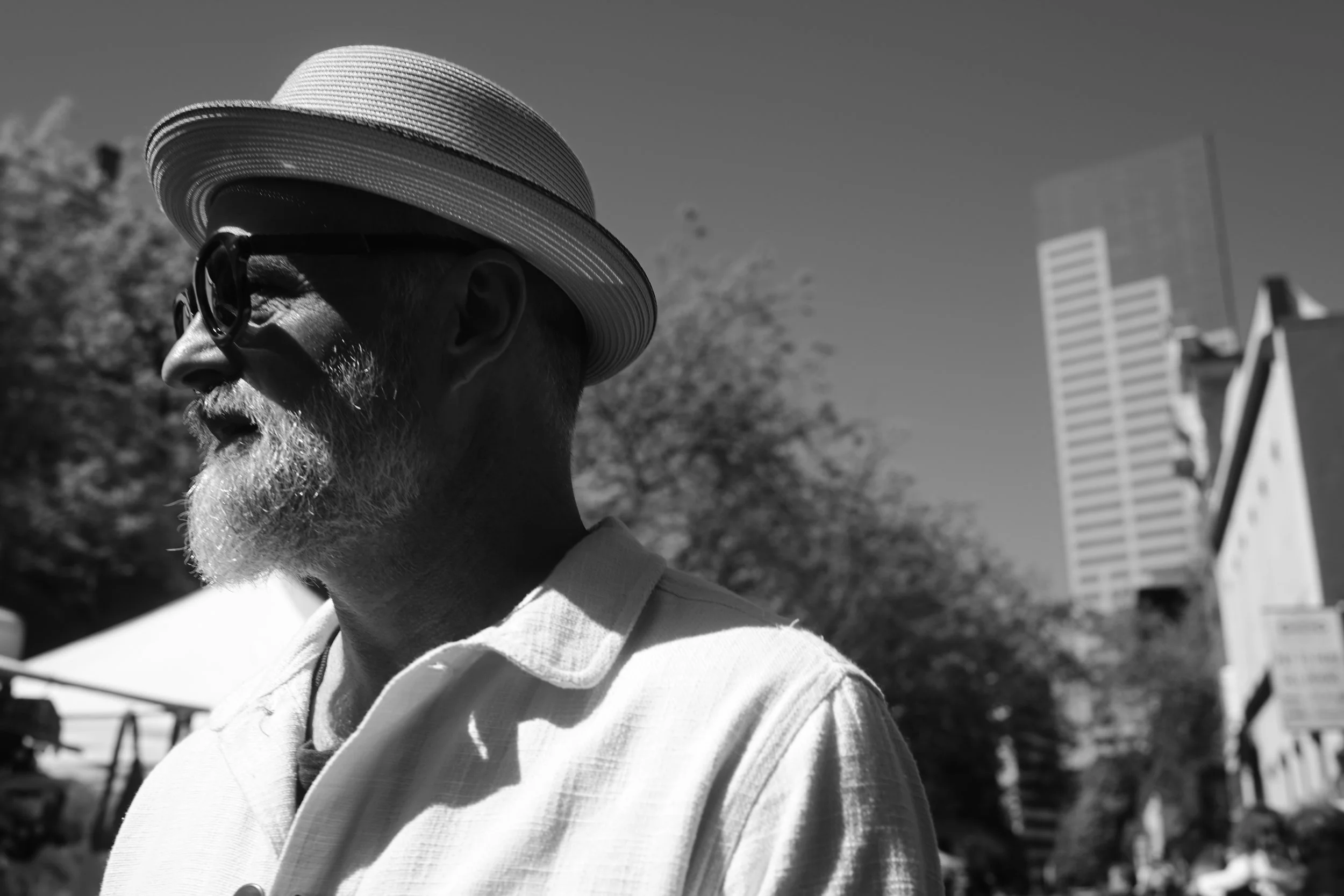 A grayscale photo of an older man with glasses and a beard, wearing a striped sunhat and a collared shirt, outdoors in a city setting with tall buildings in the background.