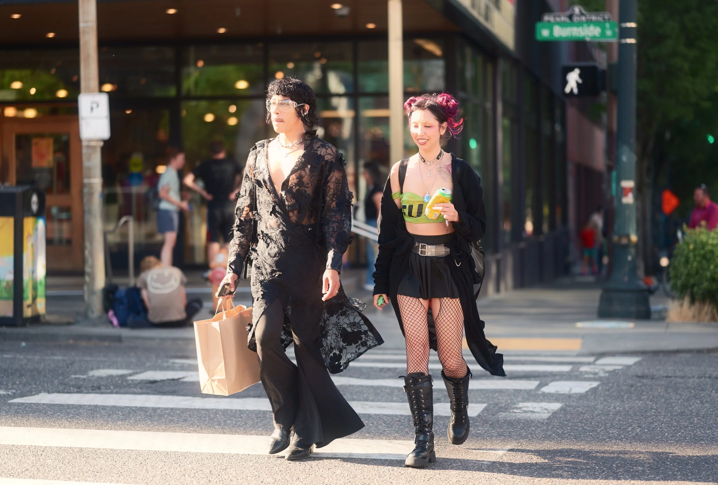 Two young women walking across a city crosswalk in the late afternoon, dressed in alternative fashion, one in a long black lace coat and the other in a black skirt, fishnet tights, and a neon green top, carrying drinks and shopping bags.