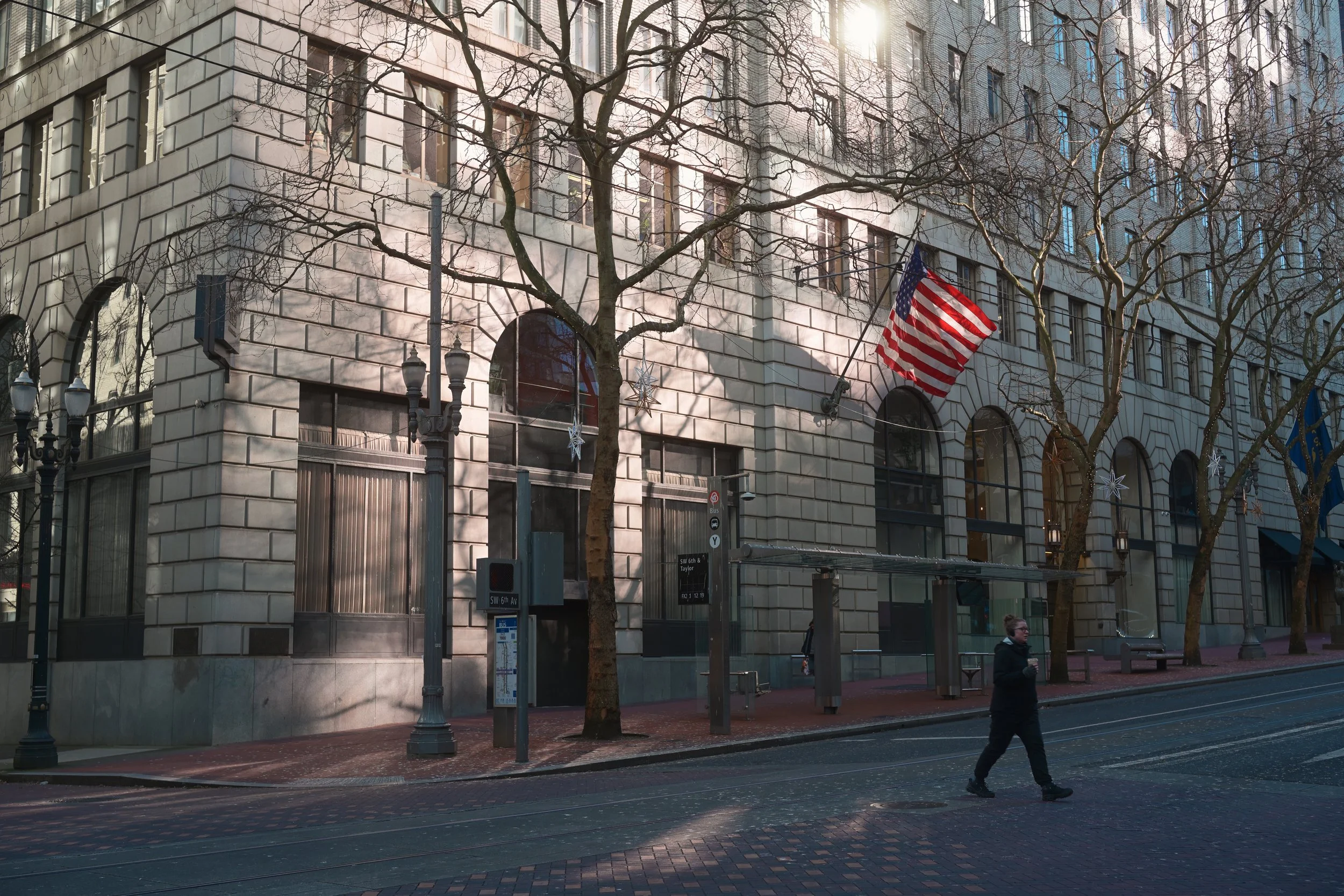 City street scene with a large beige building featuring arched windows and a flag of the United States hanging outside. Leafless trees line the sidewalk, and a woman is walking across the street. The scene is lit by sunlight, creating shadows on the 