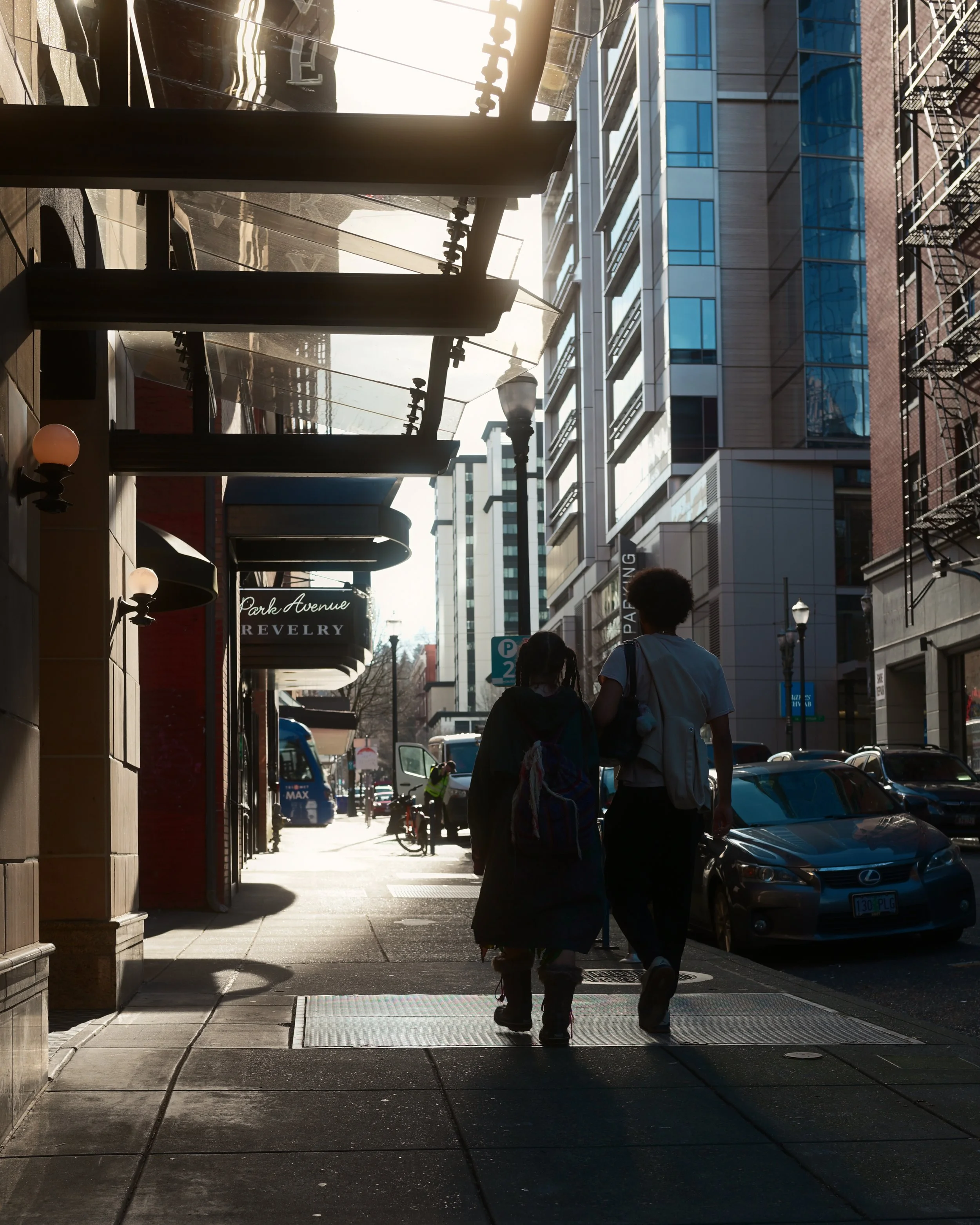 Sidewalk scene in an urban area with two people walking away from the camera, buildings with glass windows, parked cars, street lamps, and storefronts, including a jewelry store sign, under a sunlight backdrop.