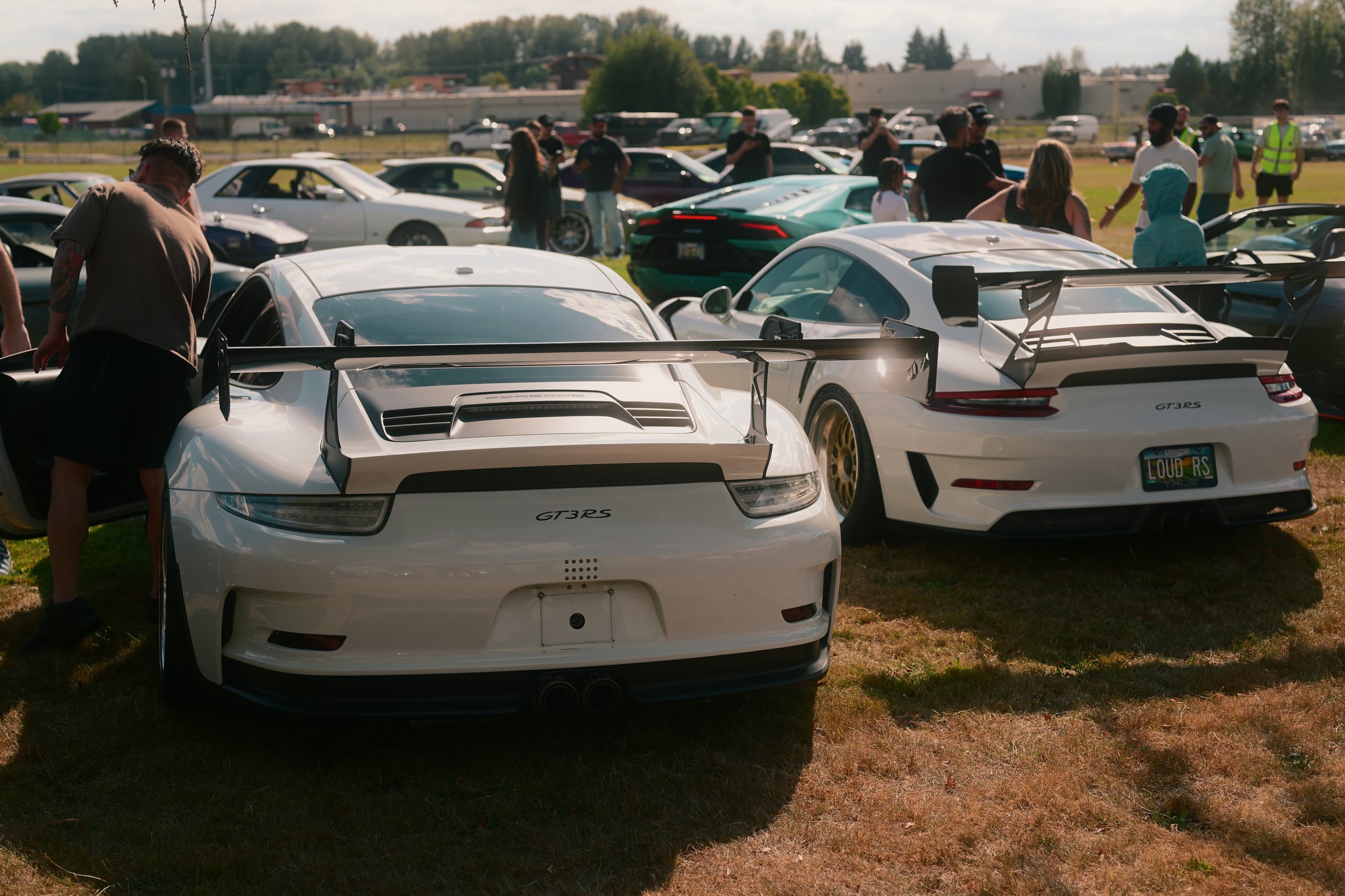 A gathering of people around several Porsche sports cars parked on a grassy field during daytime. The cars are mainly white with large rear spoilers, and the setting appears to be an outdoor car event.