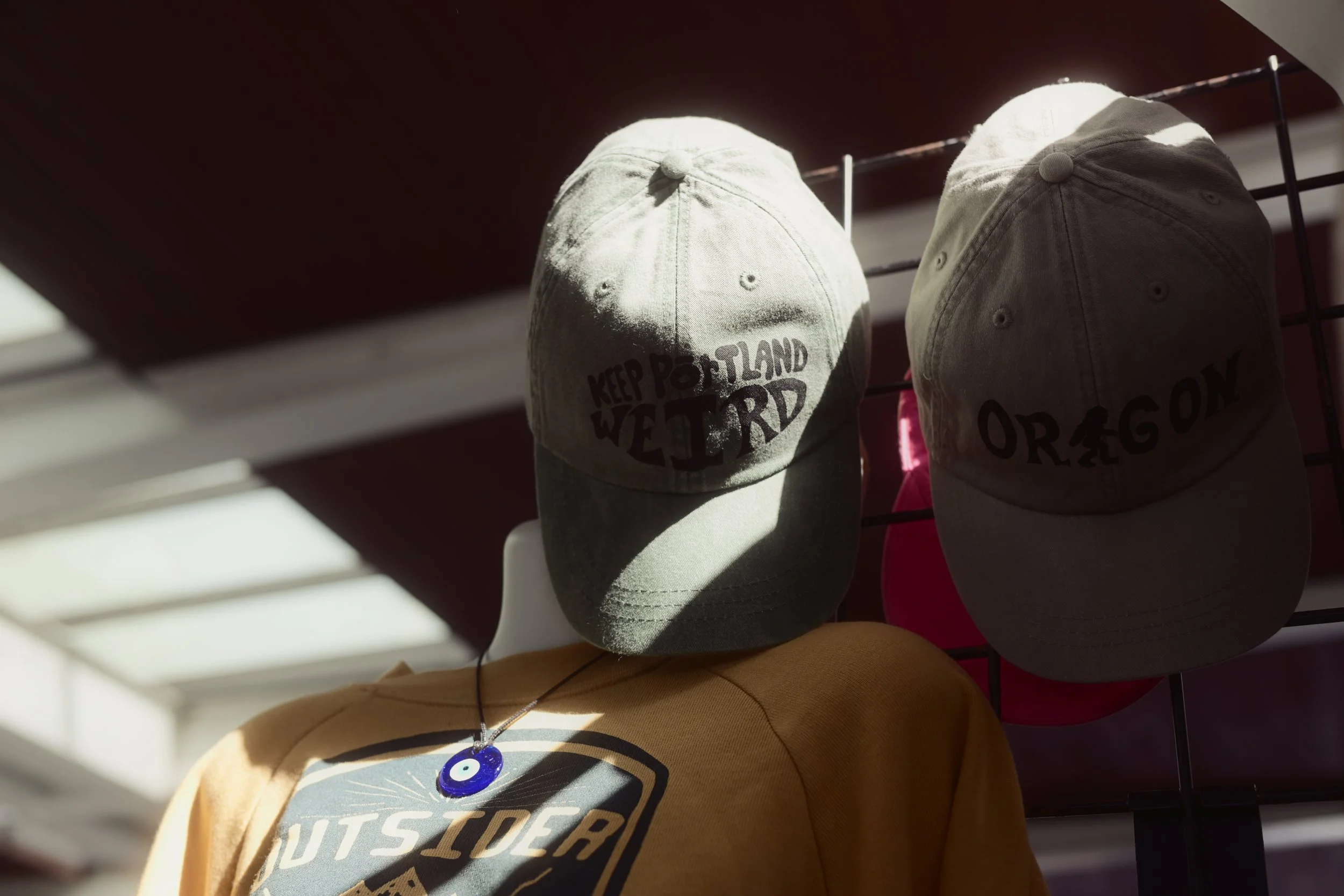Two beige baseball caps, one with the words "Keep Portland Weird" printed on it, hanging on a display rack with other hats.