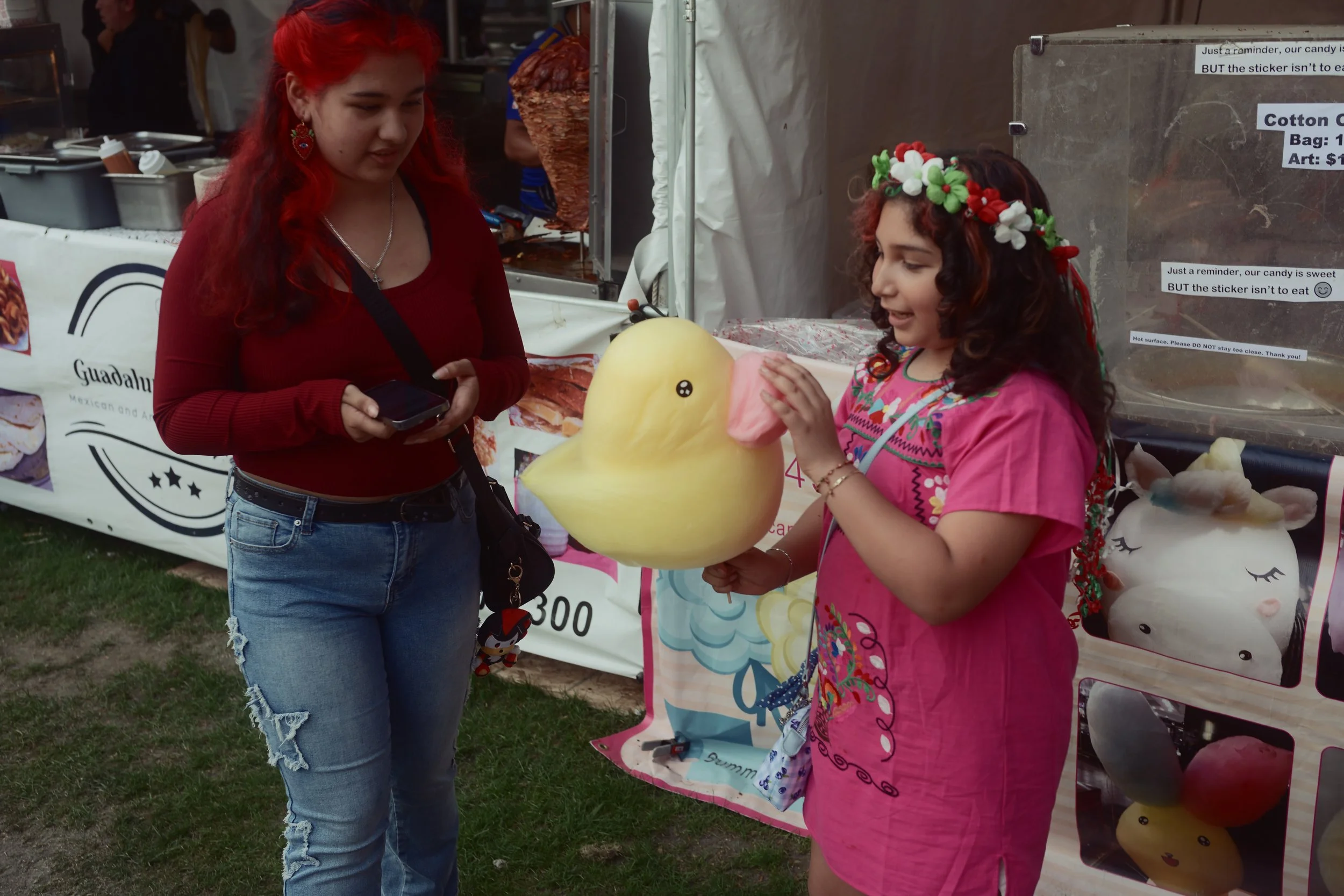 A young girl wearing a pink dress and a floral headband has a yellow duck-shaped cotton candy, and an older woman in a red top and ripped jeans is looking at her, holding a phone, at an outdoor fair or market with a food stall in the background.