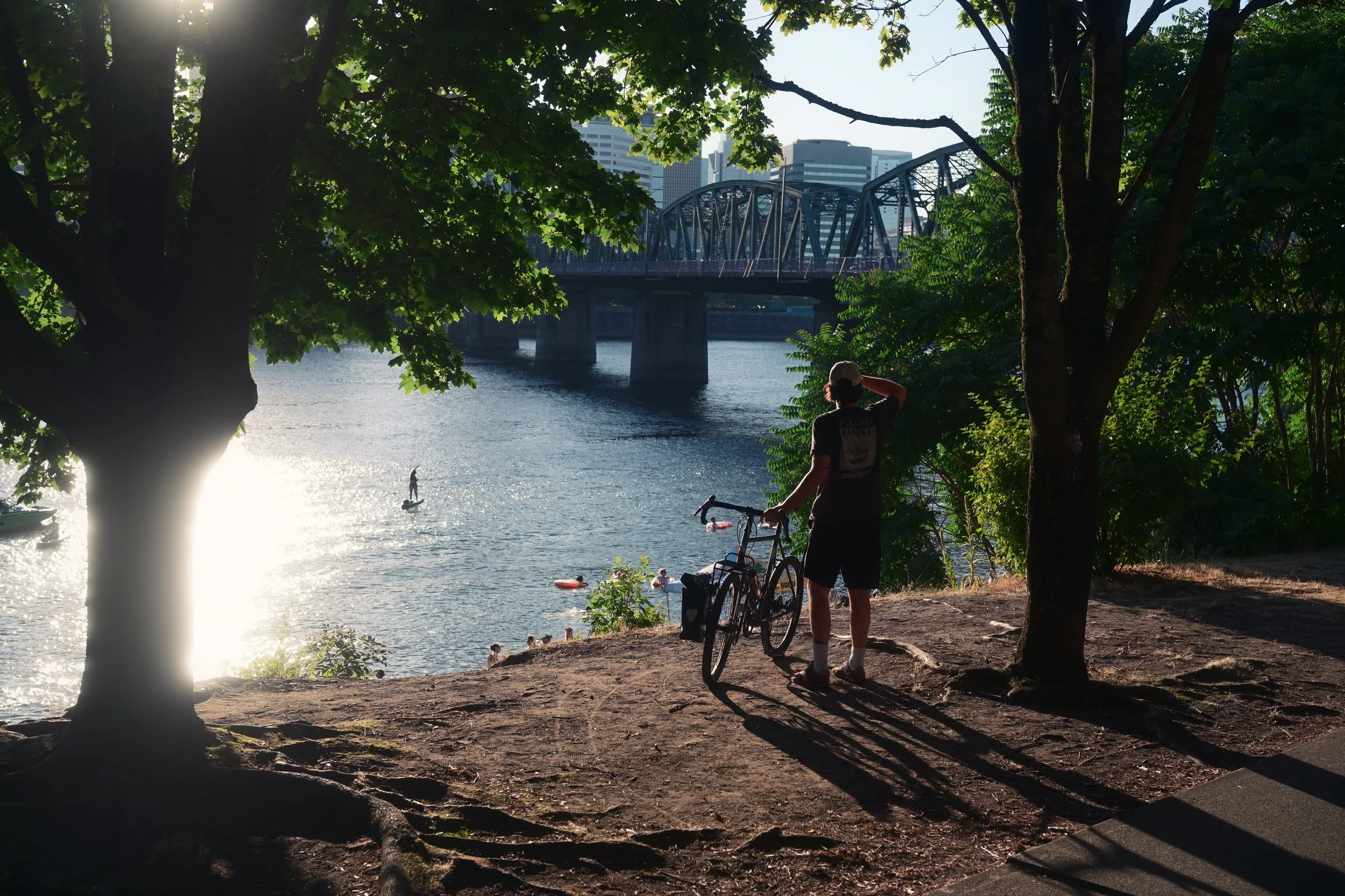 A person standing with a bicycle near the riverbank, looking at the water with a bridge and city buildings in the background, surrounded by trees and sunlight