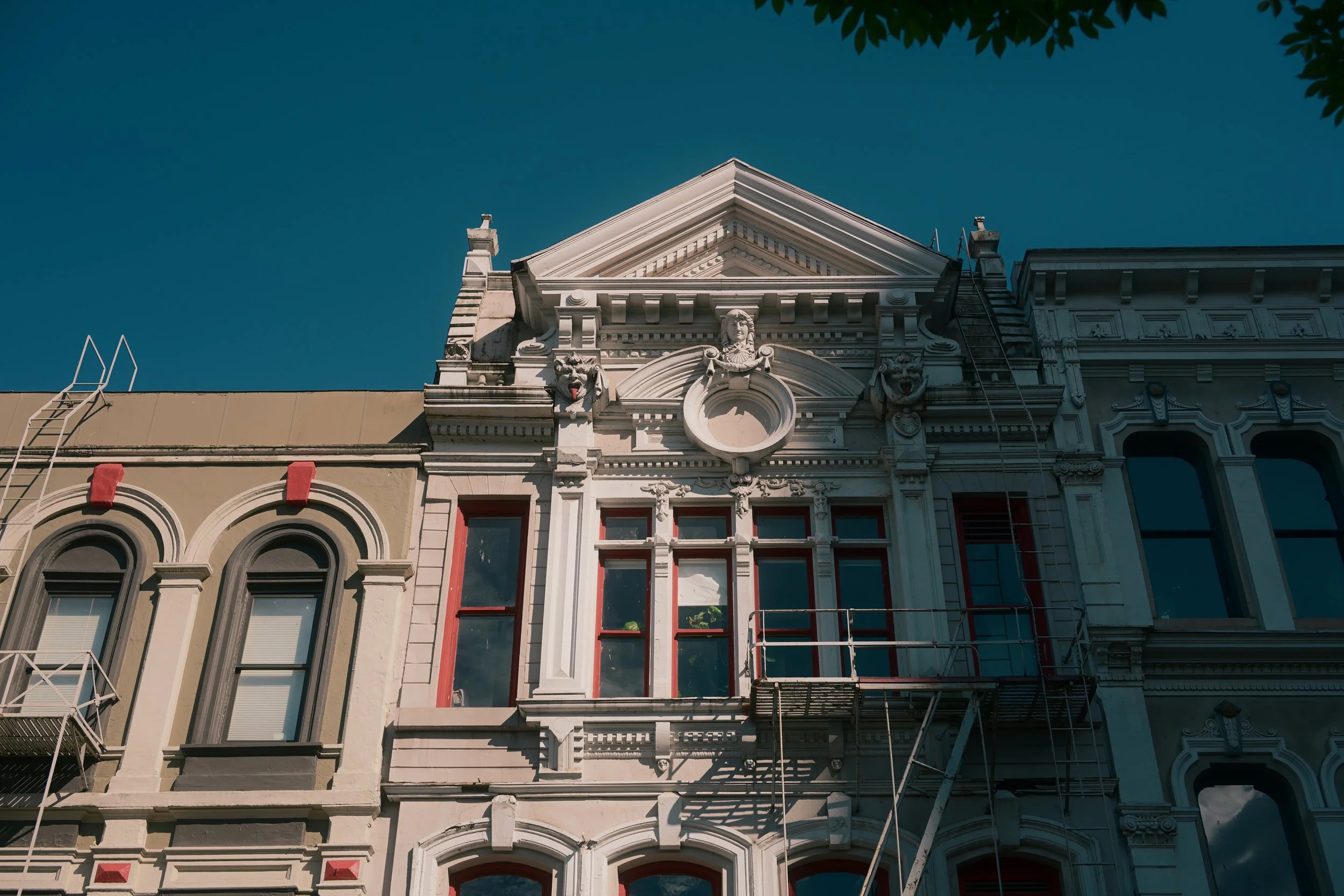 The upper part of a historic building with ornate architectural details, including crowned lion sculptures and a convex triangular pediment, against a clear blue sky. Scaffolding is present on parts of the building.