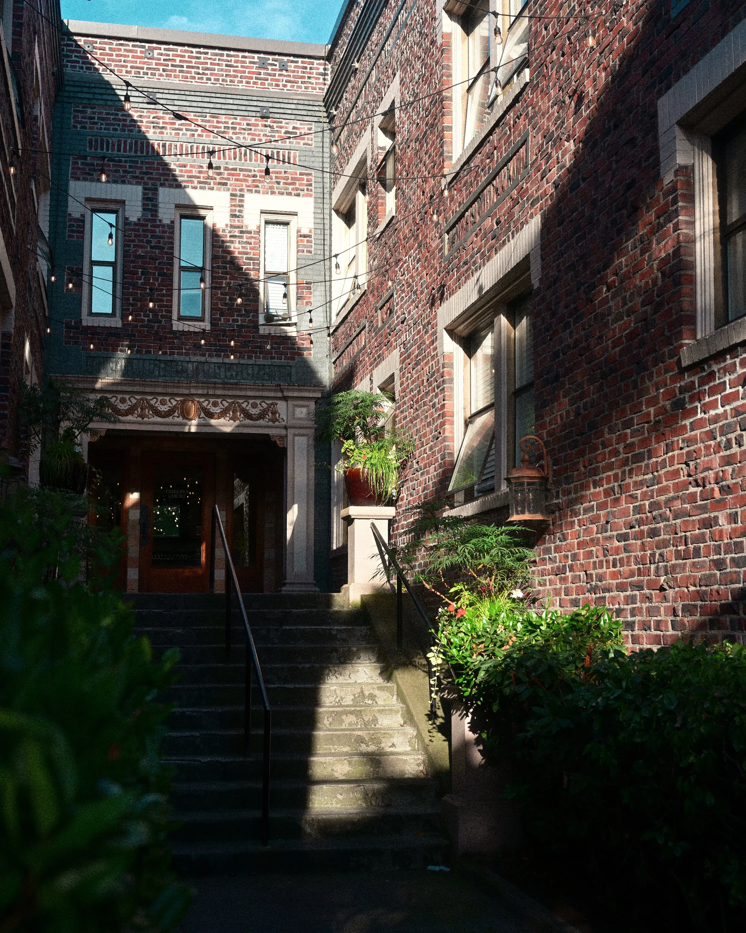 Brick courtyard with stone stairs, lush plants, windows, and string lights surrounding a historic building entrance