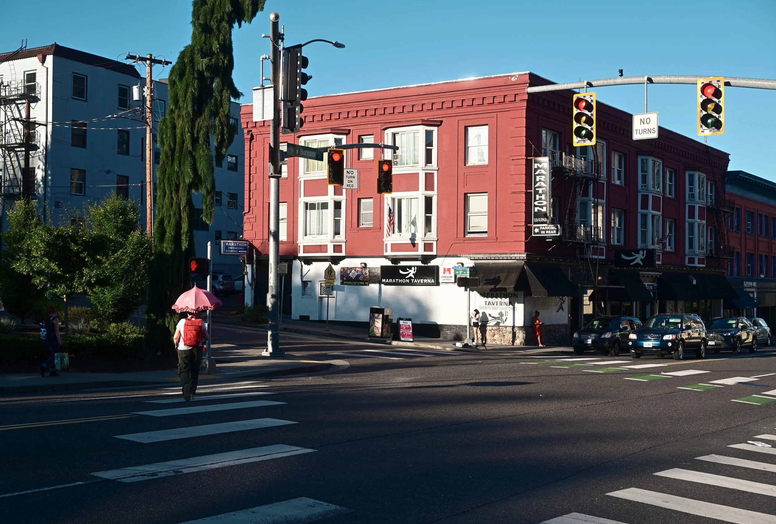 City street intersection with traffic lights, pedestrians, and a red building with a sign for Marathon Taverna.