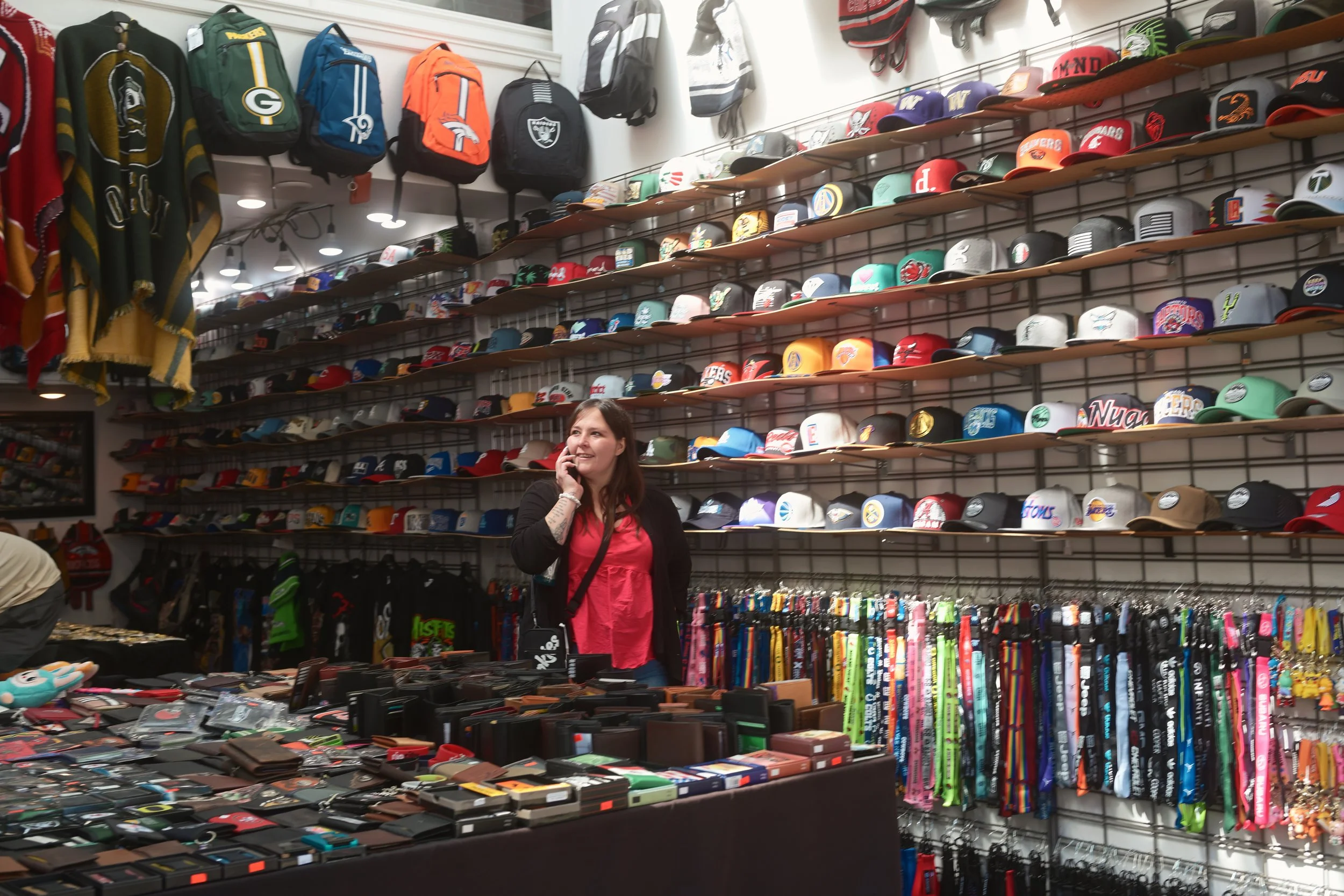 A woman shopping in a store with a large wall display of baseball caps organized on shelves behind her. The caps feature various team logos and designs.
