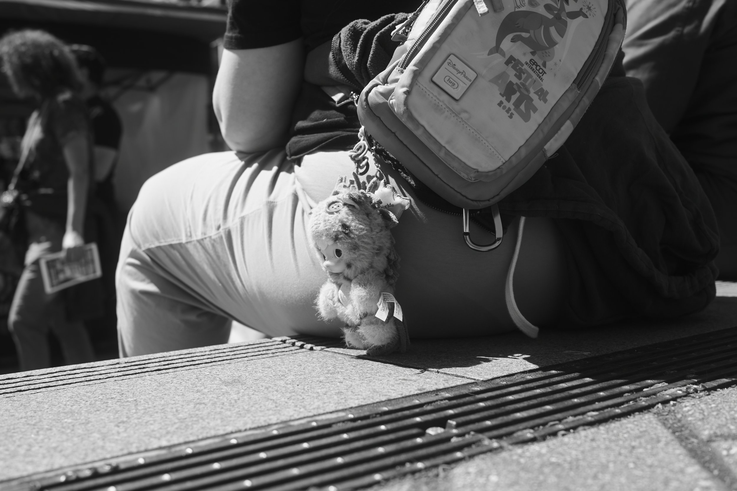 A person sitting outdoors on a bench, carrying a backpack with a Disney Sticker and holding a plush toy. There is a dog in the background.