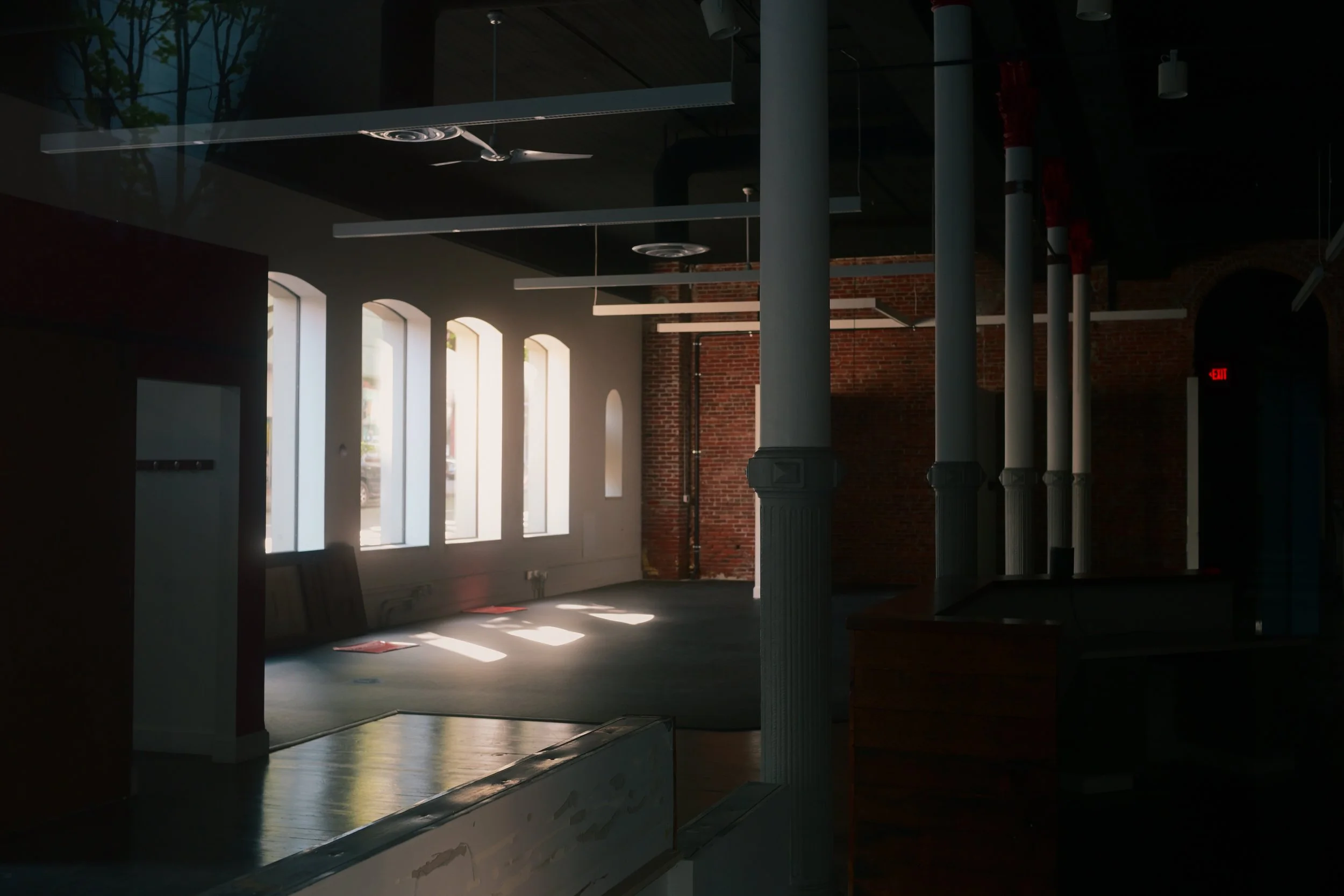 An interior space with tall, arched windows allowing natural light to illuminate the room. There are white columns and a brick wall, with hanging lighting fixtures and ceiling fans. The space appears to be an industrial-style or renovated building with exposed pipes and a mix of modern and vintage design elements.