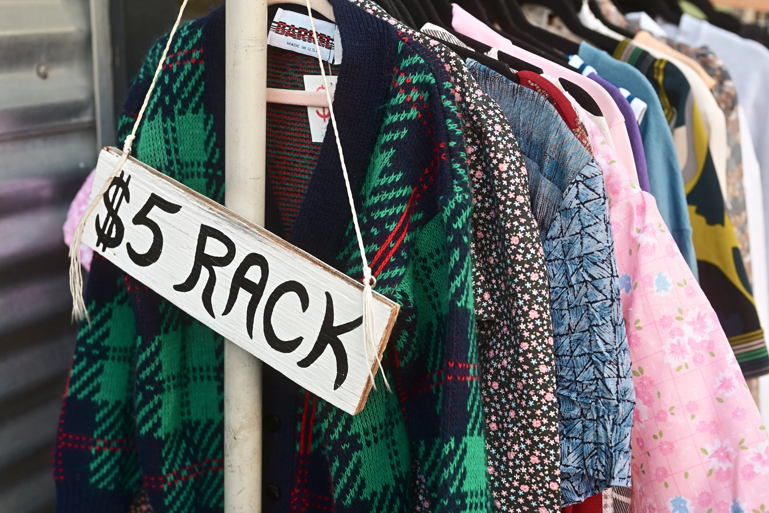 A row of sweaters and shirts hanging on a rack at a yard sale, with a sign indicating each item costs five dollars.