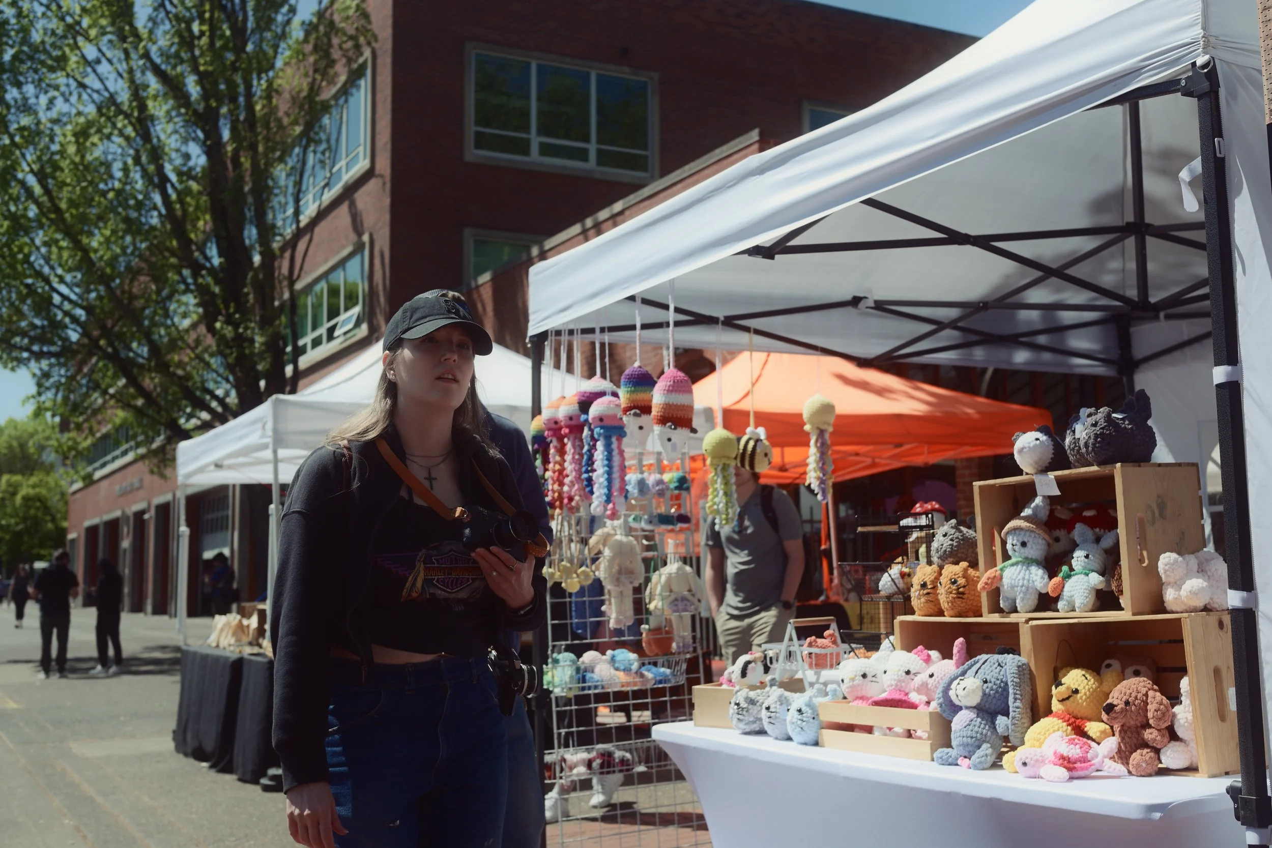 A young woman wearing a black baseball cap and dark clothing shopping at an outdoor craft market. She is holding a camera in her hand. The market has white tents and displays of handmade stuffed animals and other crafts, with trees and a building in the background on a sunny day.