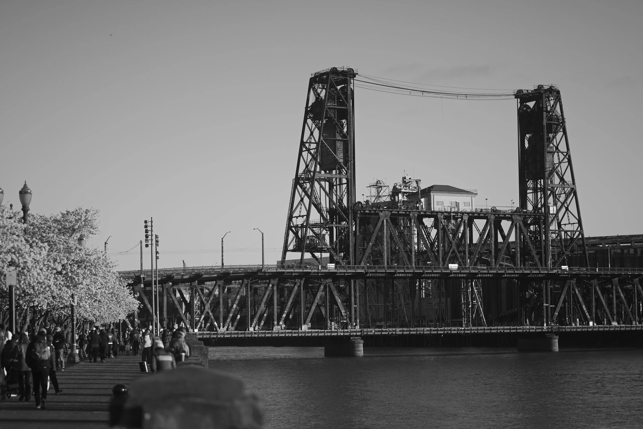 Black and white photo of a bridge with industrial towers above, with people walking along the riverside promenade.