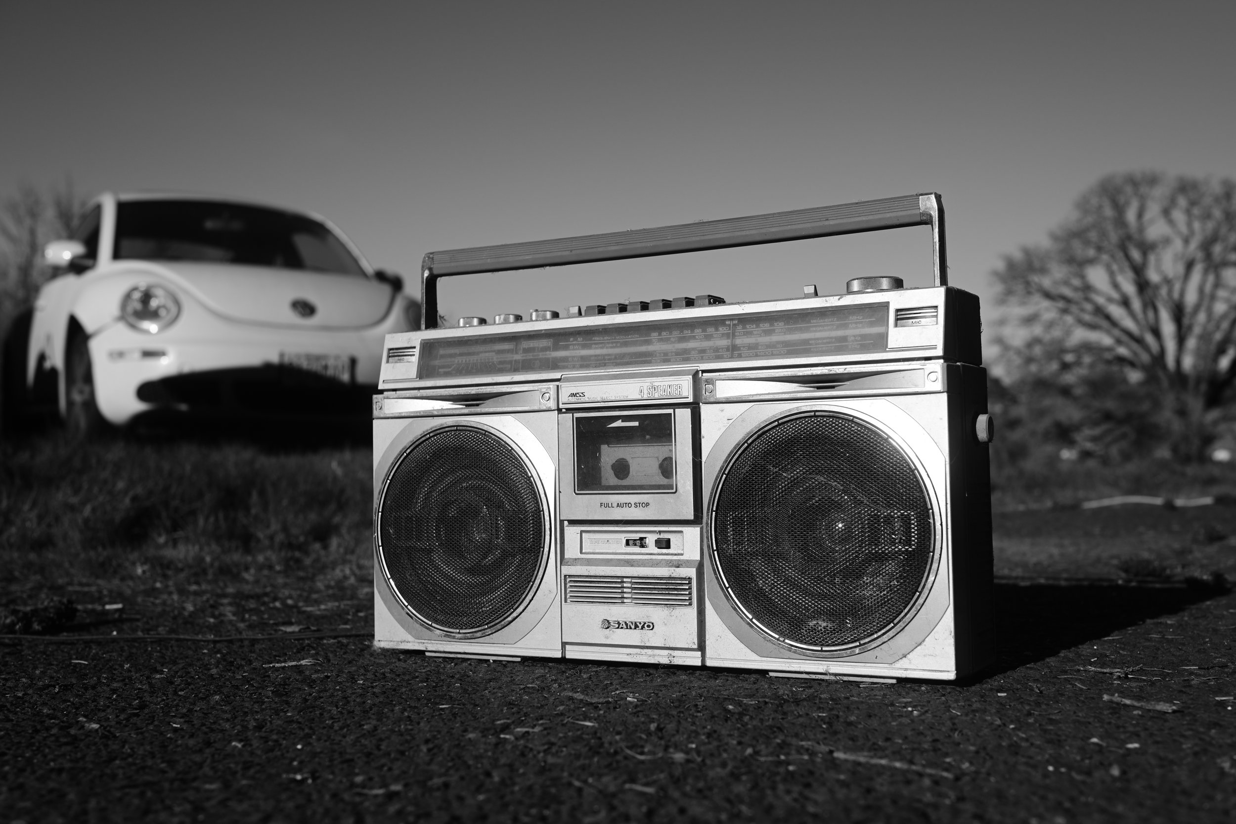 A vintage portable stereo with speakers and a radio, in front of a blurred car and trees in the background, in a black and white photo.
