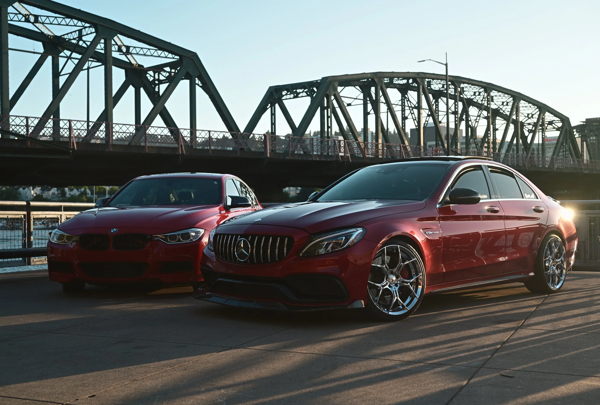 Two red luxury cars, a BMW and a Mercedes-Benz, parked on a city street at sunset with a steel bridge in the background.