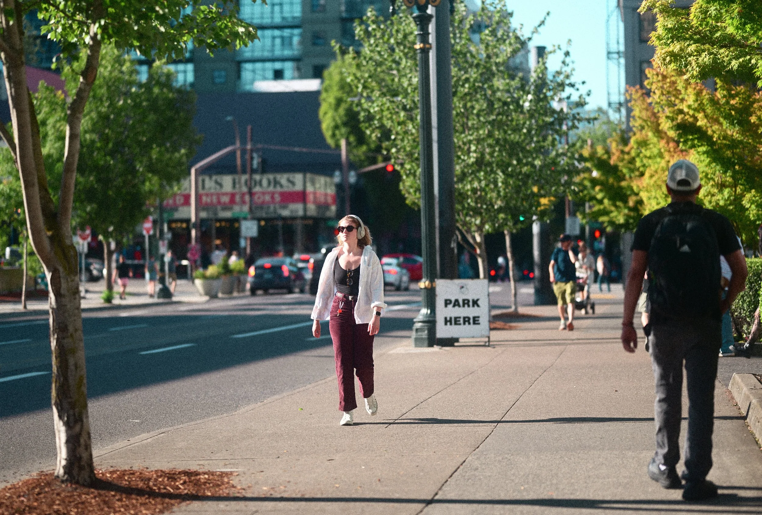 People walking on a tree-lined city sidewalk beside a busy street, with a woman wearing headphones and a park-here sign.