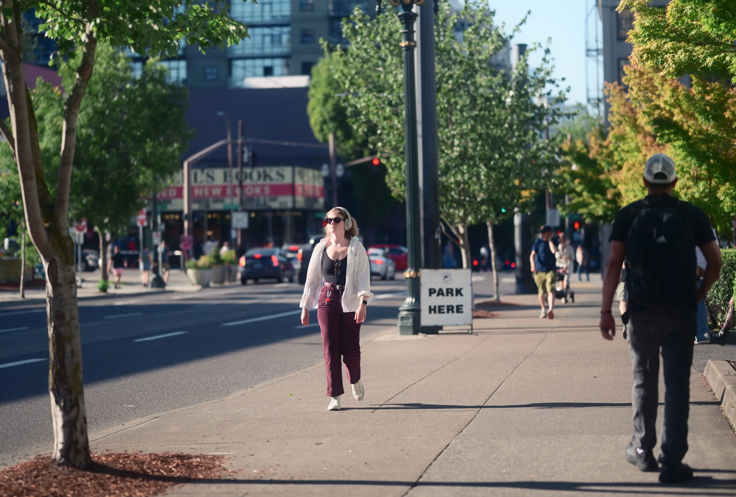 People walking on a city sidewalk, trees lining the street, cars on the road, a woman with headphones and sunglasses walking towards the camera, a man with a backpack walking away, and a sign that says 'Park Here' on a post.