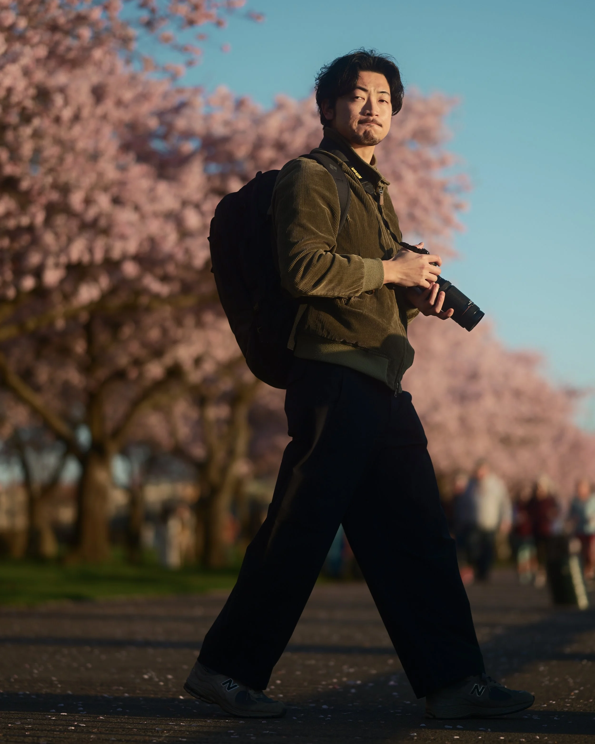 A man walking outdoors holding a camera, with pink cherry blossom trees in the background during sunset.
