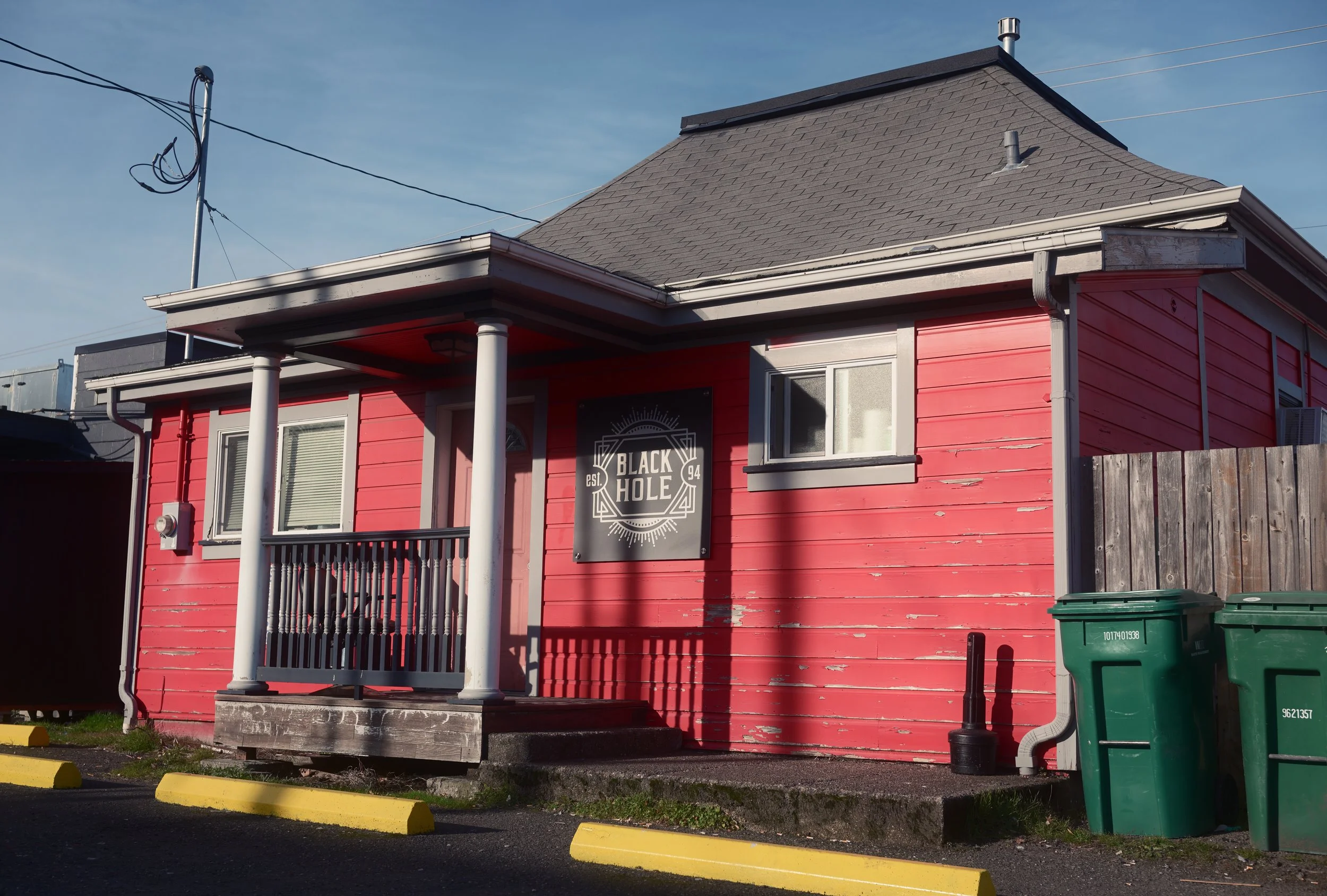 A pink wooden building with a small porch supported by two white columns, a black sign that reads 'Black Hole' on the front, and two green trash bins to the right. The building has a grey shingle roof and a small window on the right side.