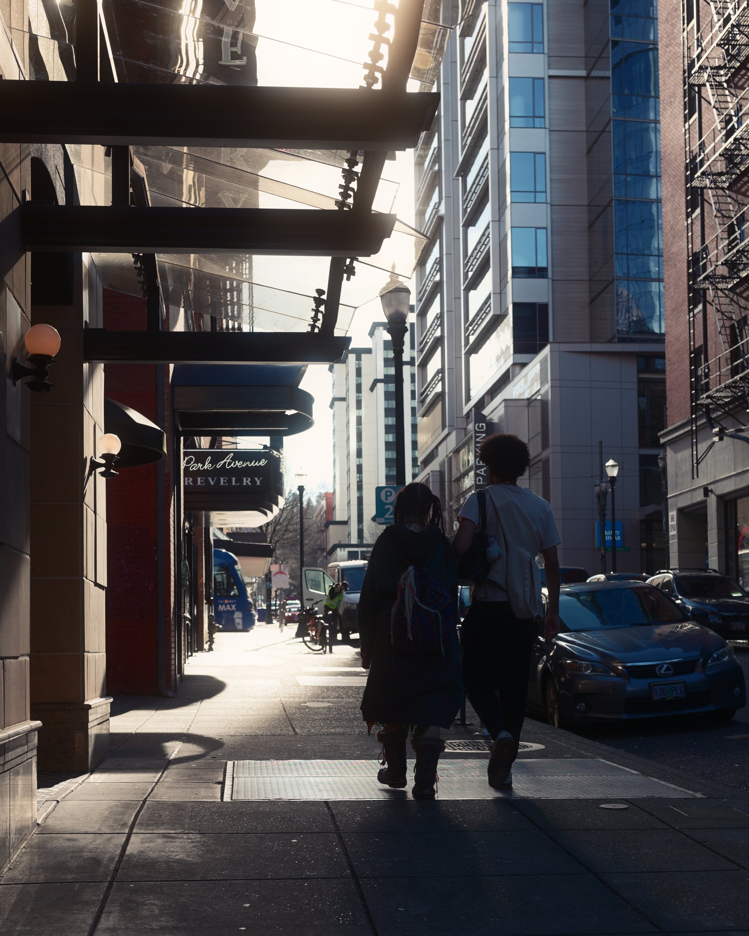 City street scene with two pedestrians walking on sidewalk, tall modern buildings, parked cars, street lamps, and a store with a sign that reads 'Park Avenue Jewelry'.