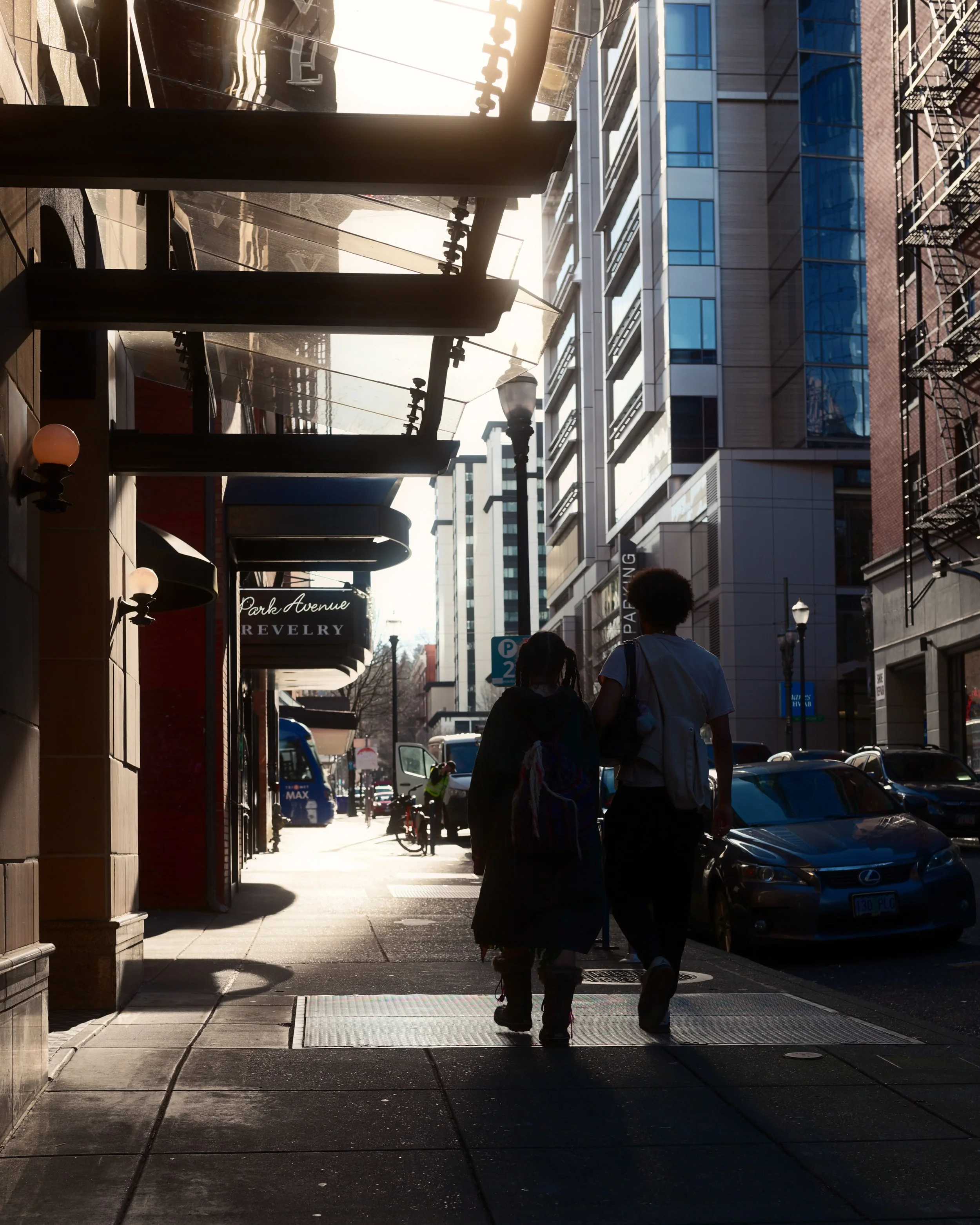 Two people walking on a city sidewalk in the late afternoon, surrounded by tall buildings with reflective windows, parked cars, and a streetlamp.