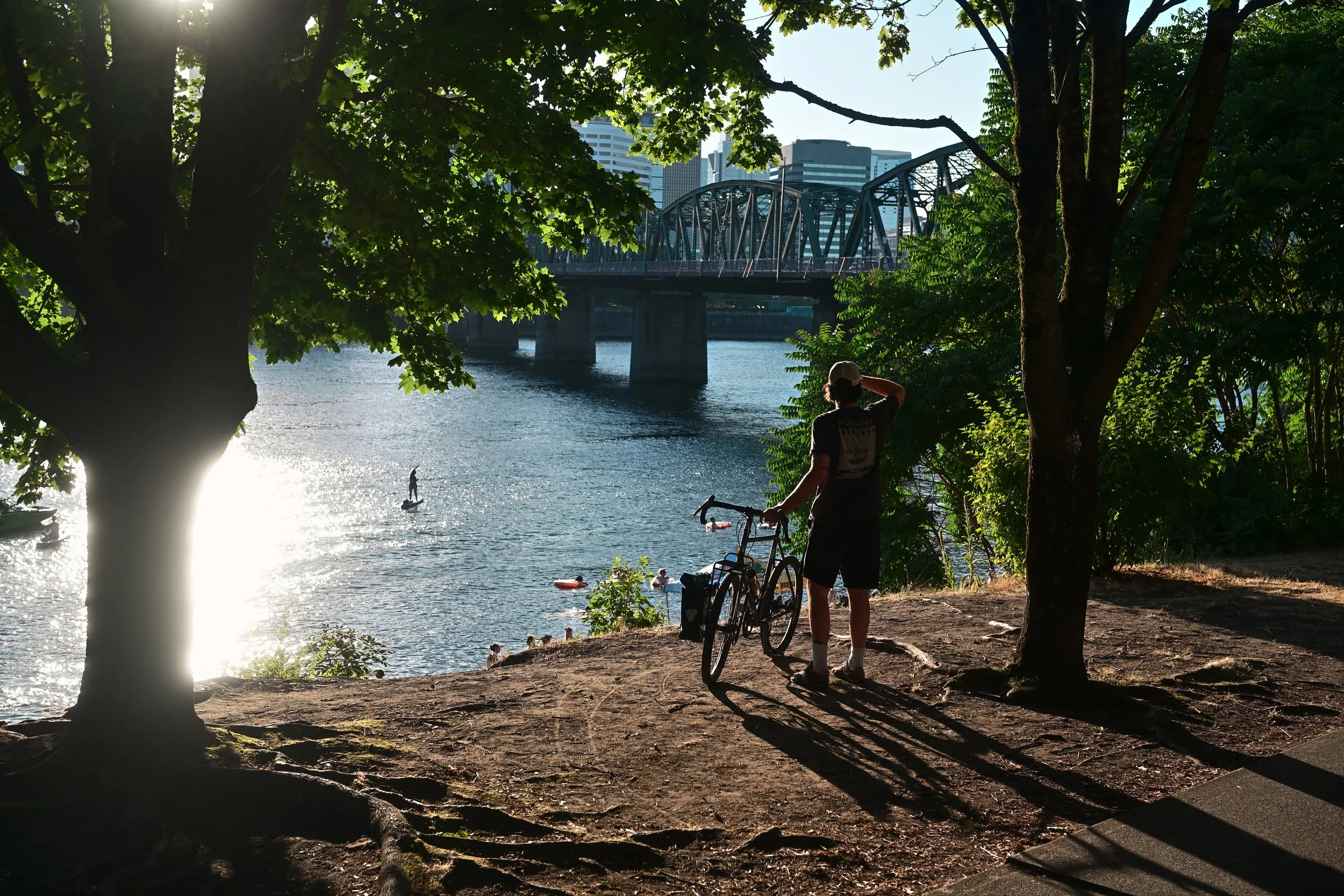 A person with a bicycle next to the water, looking at paddleboarders, under trees with a cityscape and bridge in the background.